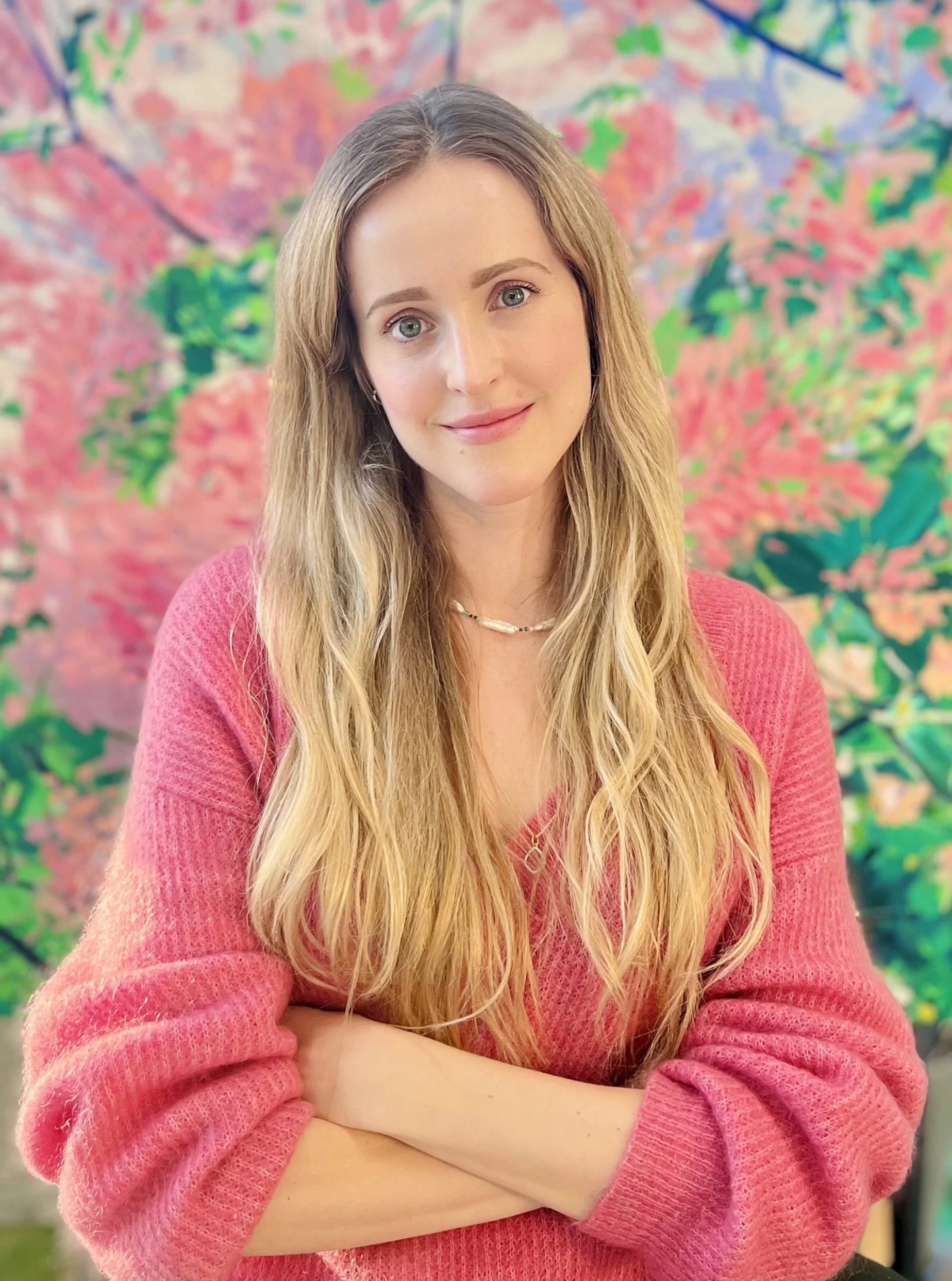 A young woman with long blonde hair and blue eyes, wearing a pink sweater and a necklace, smiling and crossing her arms in front of a colorful, abstract floral background.