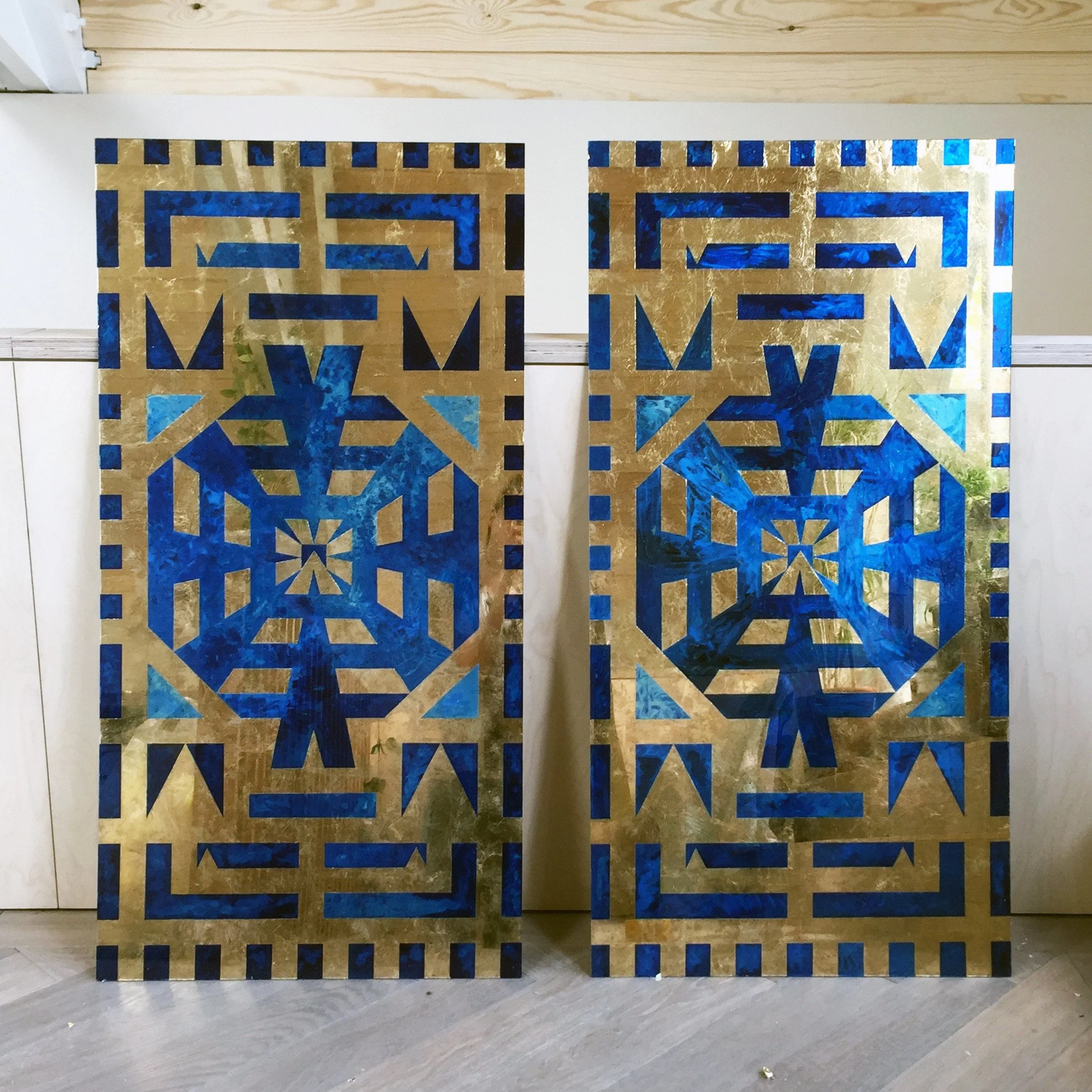 Decorative gilded panels with geometric blue and gold patterns leaning against a white wall and wooden ceiling.