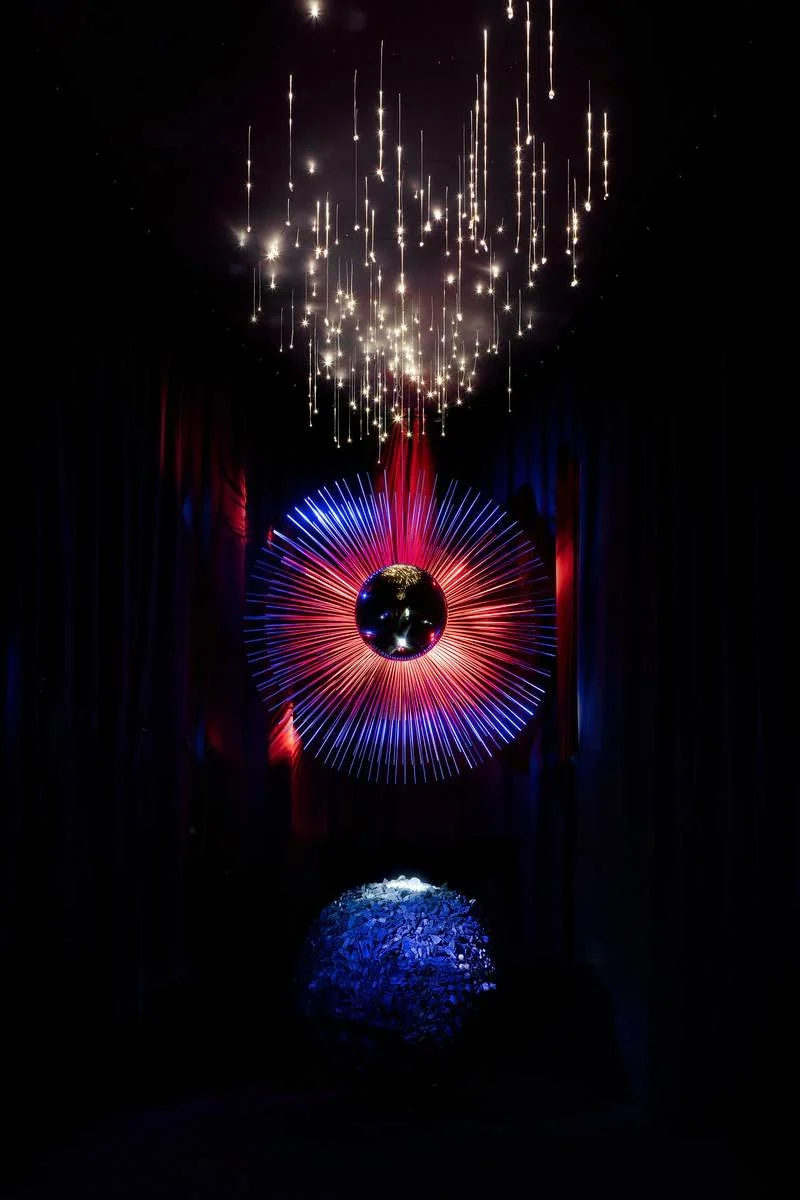 A dark room with hanging string lights, a colorful firework display in the background, and a reflective orb on the floor, representing the North Star.