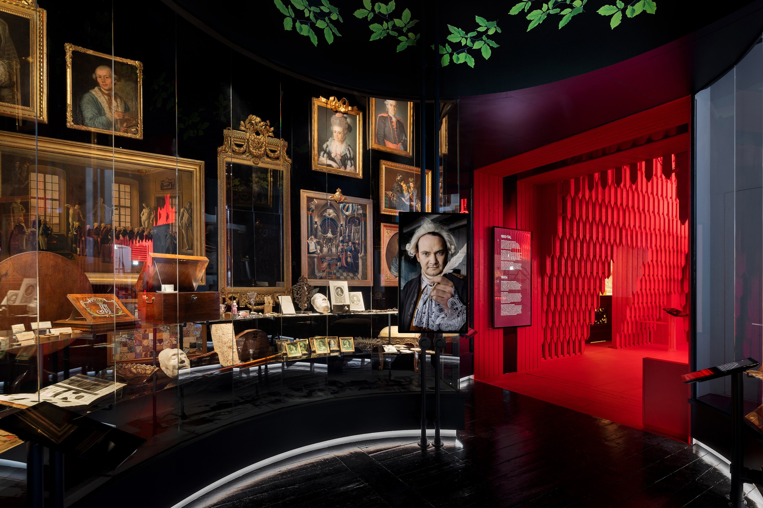 A museum exhibit with framed portraits and artifacts. A person is taking a selfie in front of the display, which includes a portrait of a woman holding a book, a portrait of a man in a military uniform, and other classical paintings. The exhibit spac