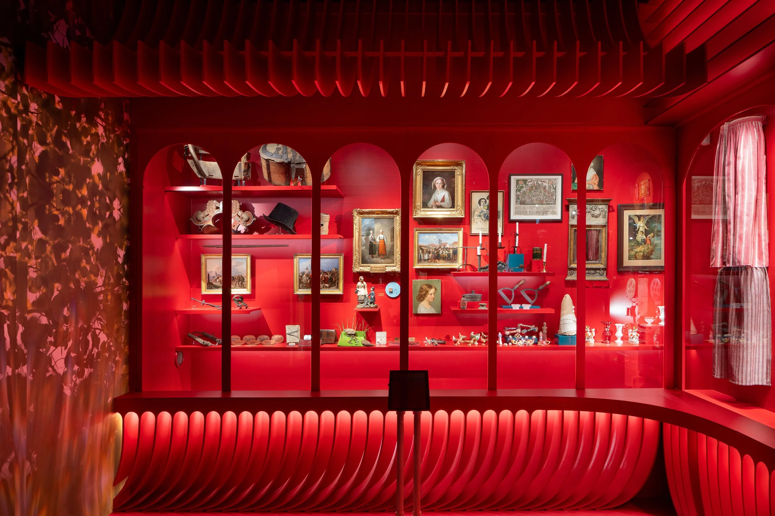 Red display case in a museum with various historical artifacts including paintings, sculptures, and jewelry, with a red background and arch-shaped dividers.
