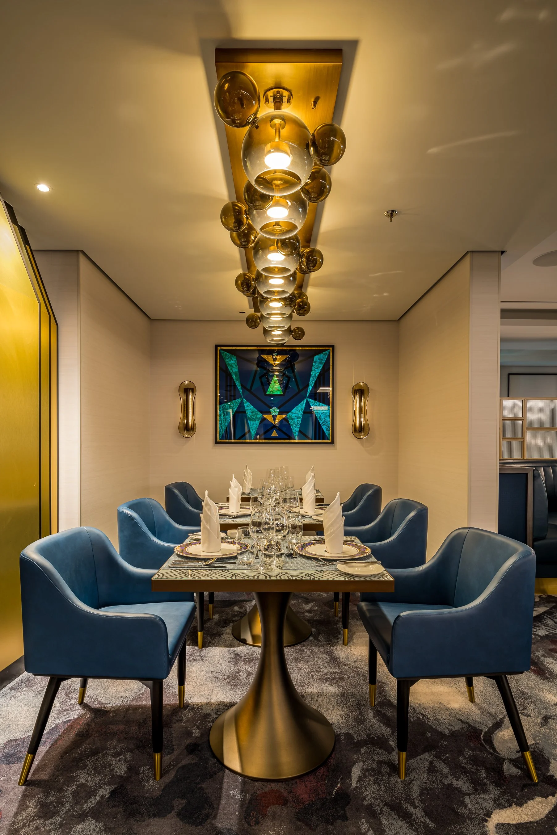 Luxury dining room with a long table, dark gray patterned tablecloth, white napkins, glassware, and blue velvet chairs with gold accents. Modern abstract artwork on the wall, gold wall sconces, and a contemporary chandelier hanging from a gold ceilin