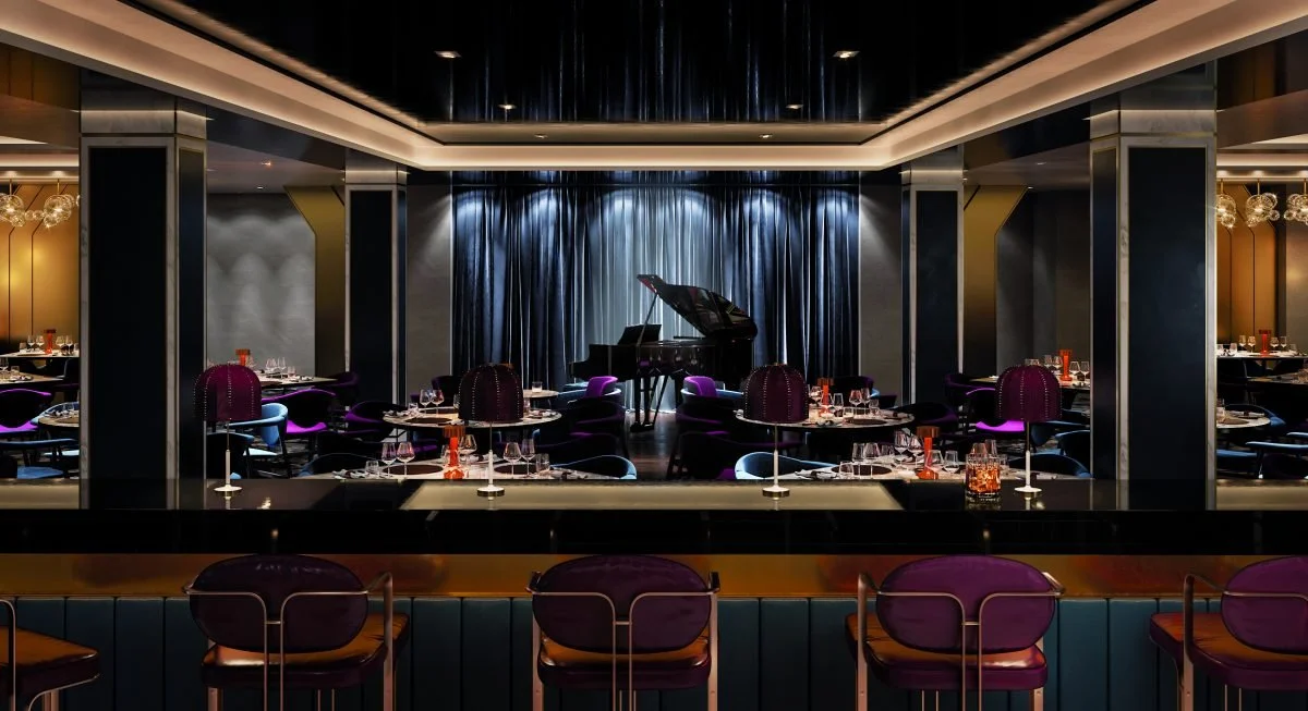 Upscale restaurant with a piano on stage, surrounded by tables with purple chairs and elegant place settings, dim lighting, and dark curtains in the background.