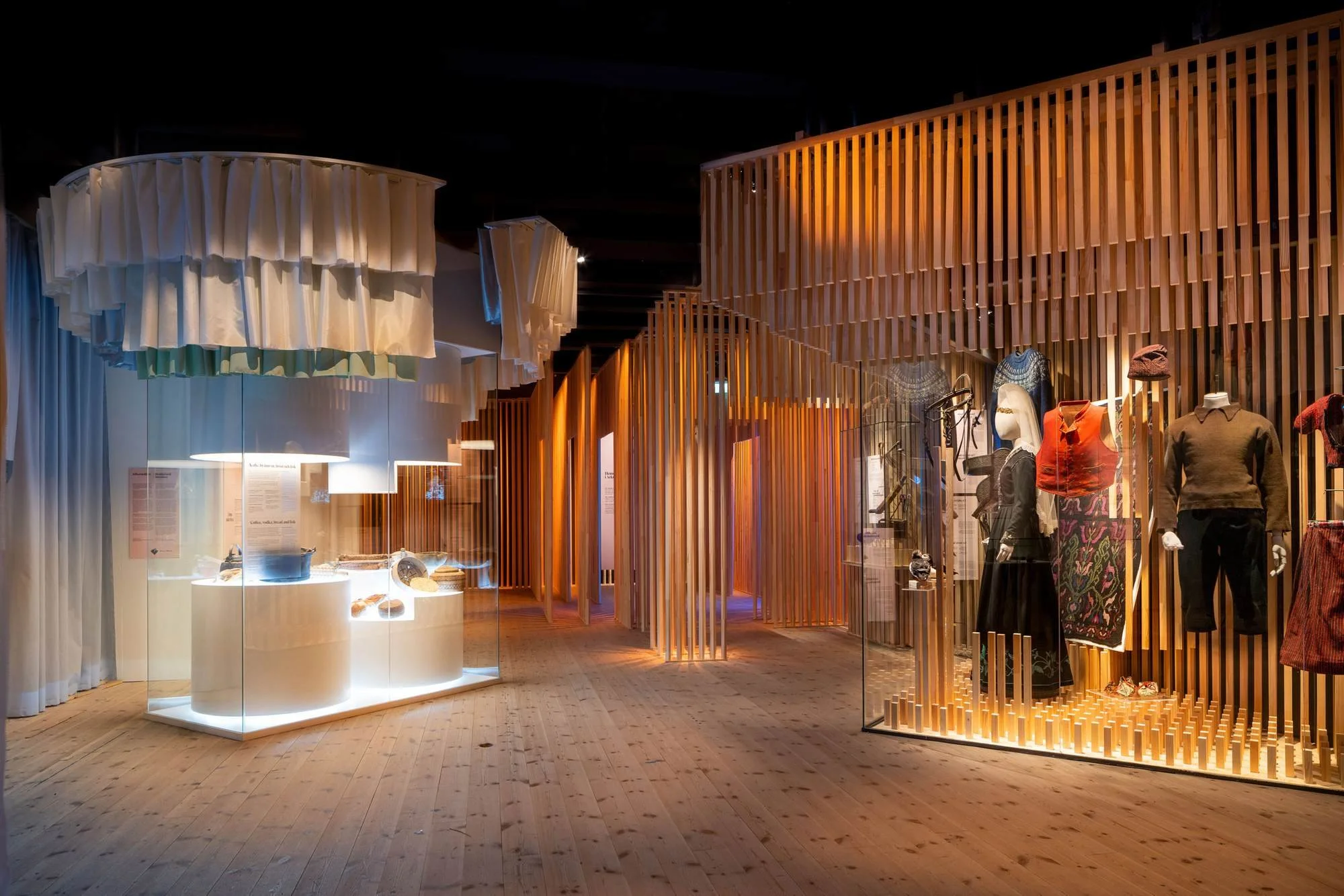 Interior of a museum with wooden and fabric display structures, mannequins dressed in clothing, and glass display cases illuminated from within.