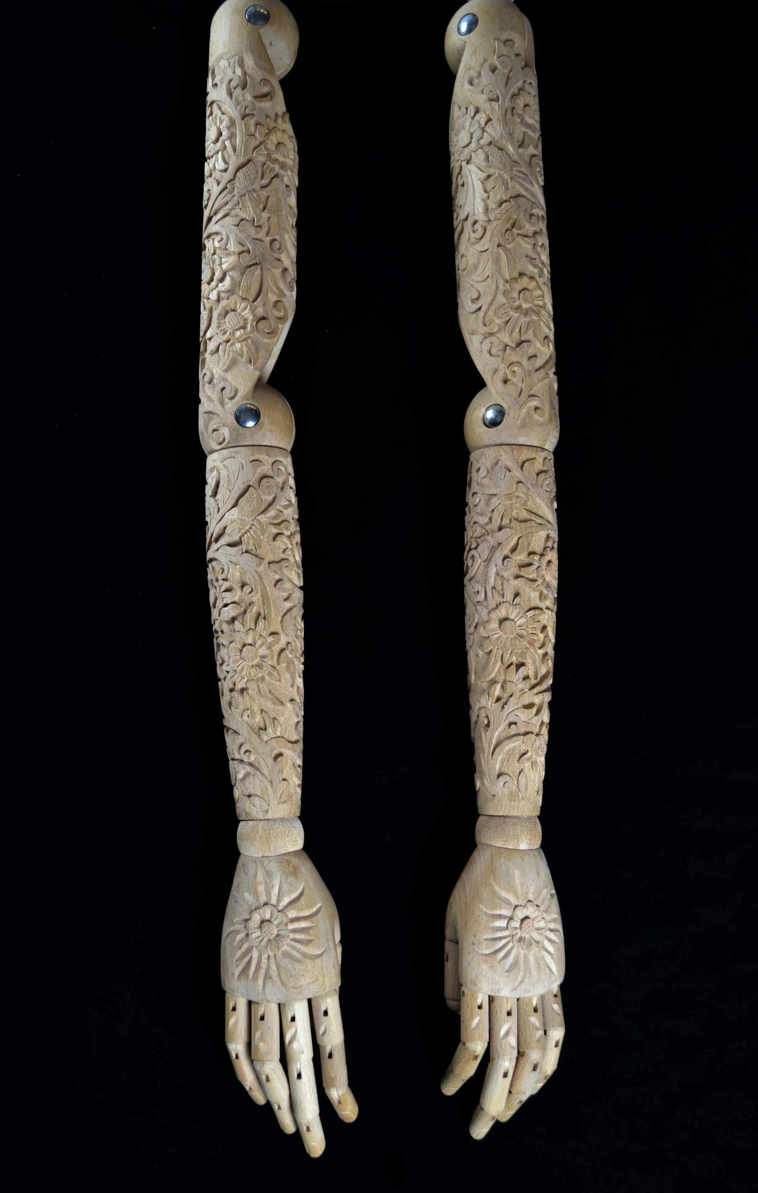 Decorative wooden, mannequin hands with hand-carved floral patterns and metal fasteners on a black surface.