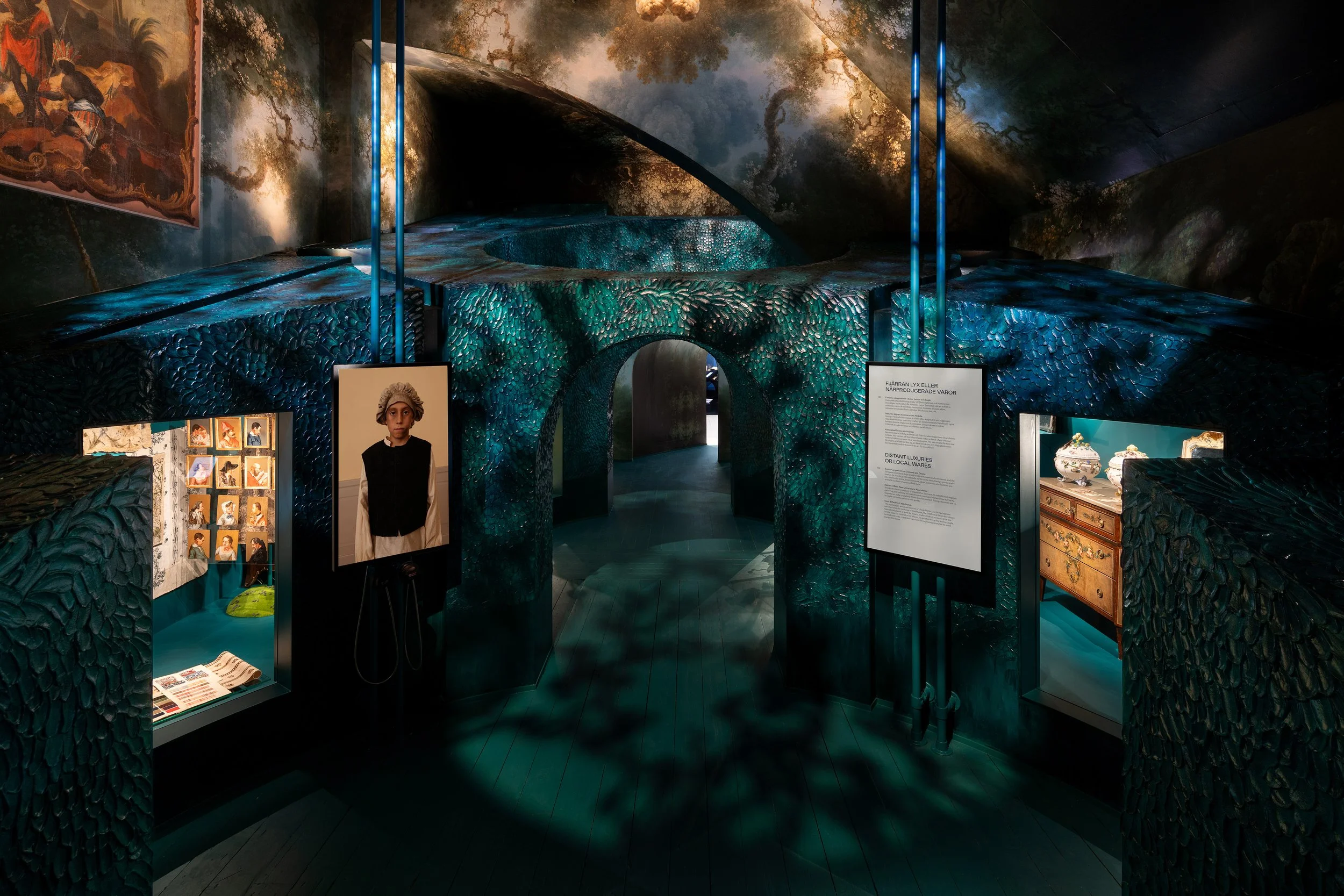 An indoor exhibit featuring a structure that resembles a topiary garden with textured leaf surface walls, in teal and blue shades, leading to an archway. The ceiling has a mural of a misty forest scene.