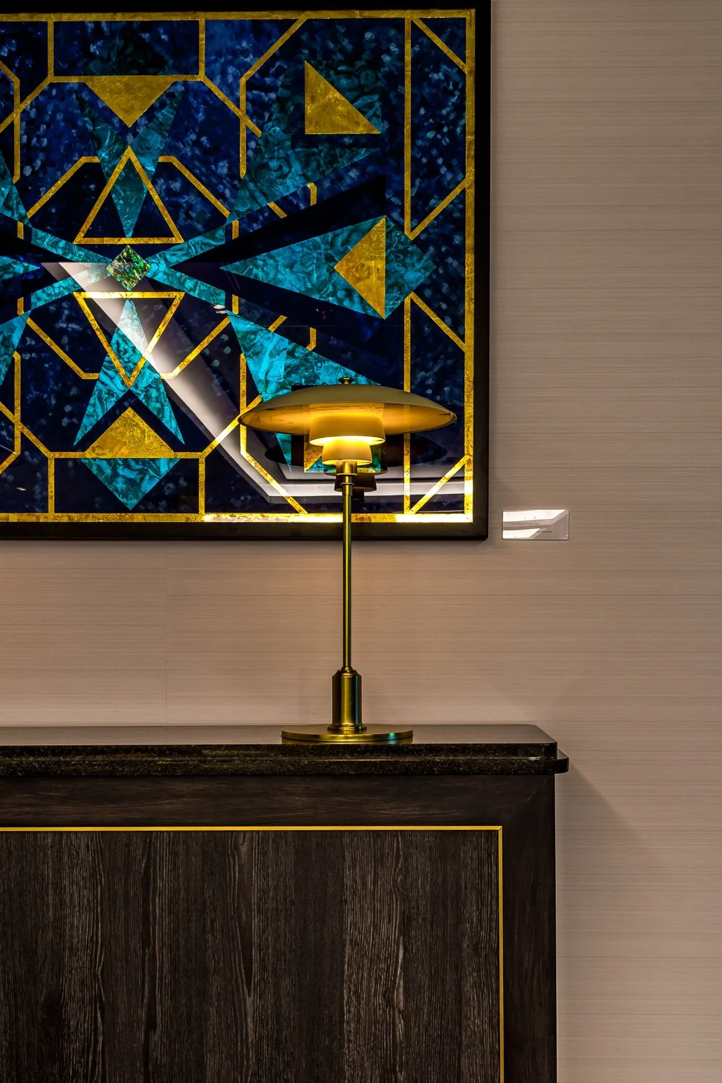 A gold-colored table lamp with a round shade on a wooden cabinet in front of a large geometric gilded glass art piece with blue, green, and gold patterns.