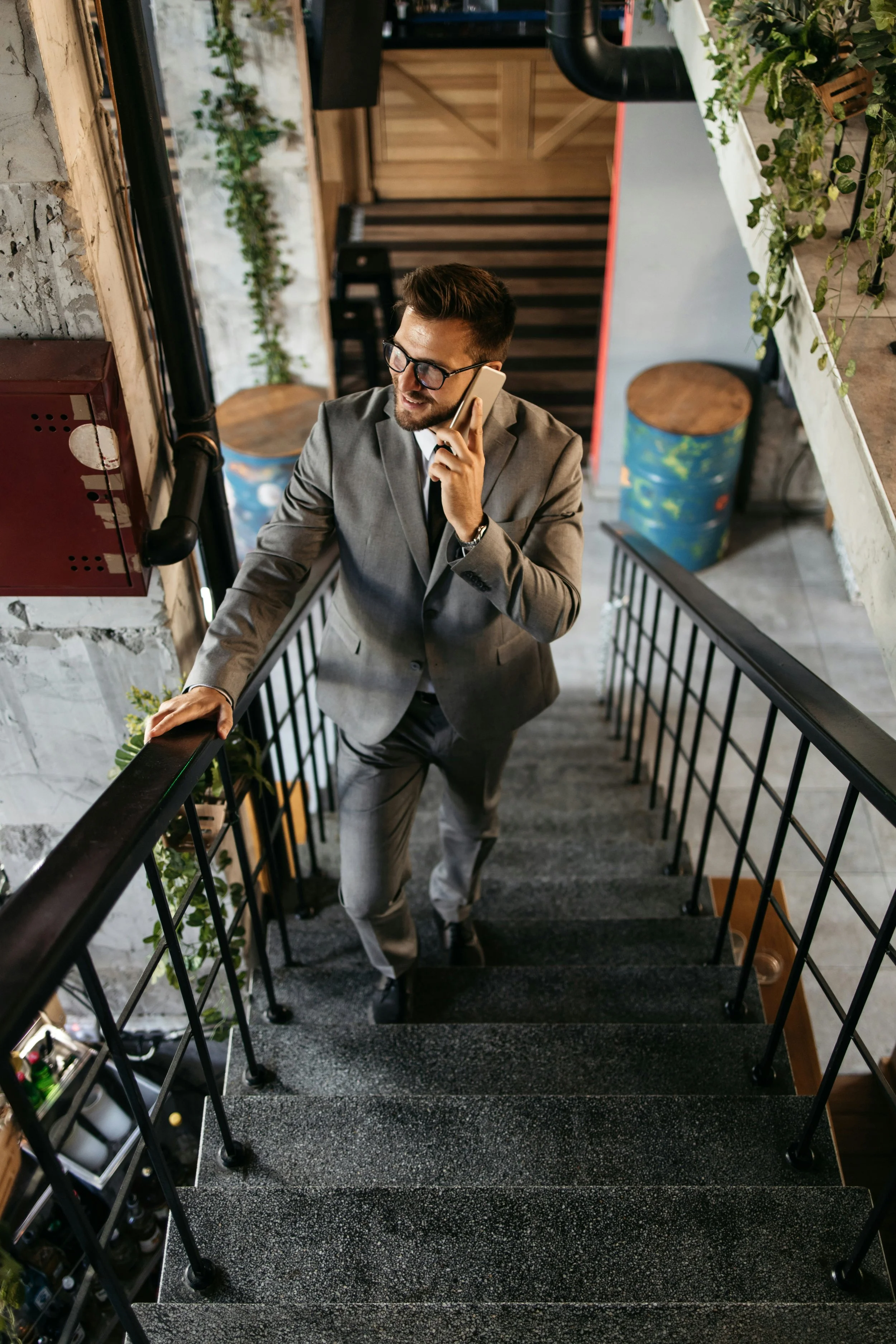 A man in a gray suit with a white shirt and black tie, wearing glasses, is smiling and talking on a cell phone while descending a black staircase with a metal railing in a modern building with exposed brick and green plants.