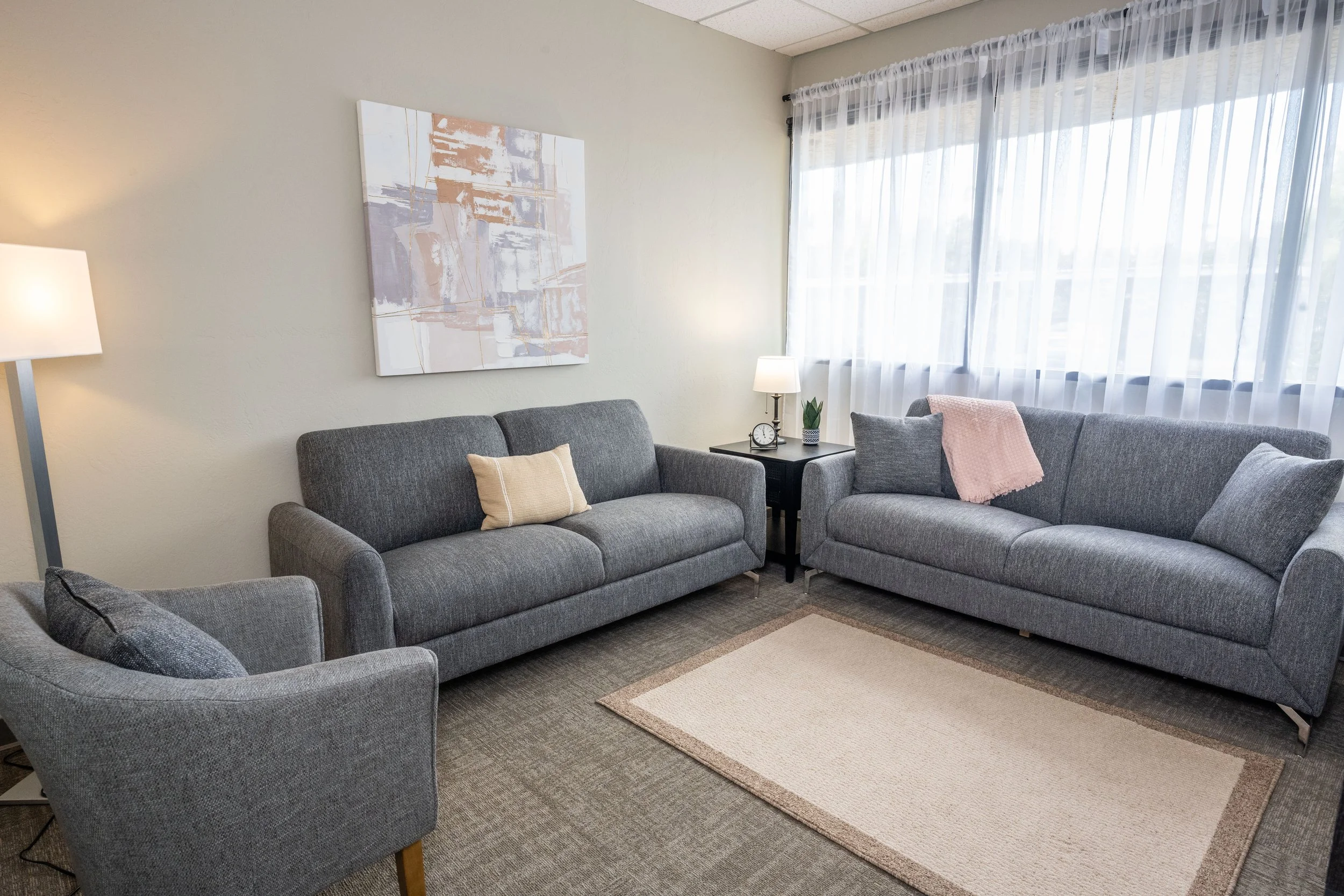 Therapy room with two gray sofas, a black side table with a lamp and clock, abstract wall art, sheer curtains, and beige rug.