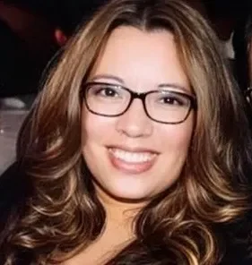 A young woman with light brown, wavy hair, glasses, and a bright smile.