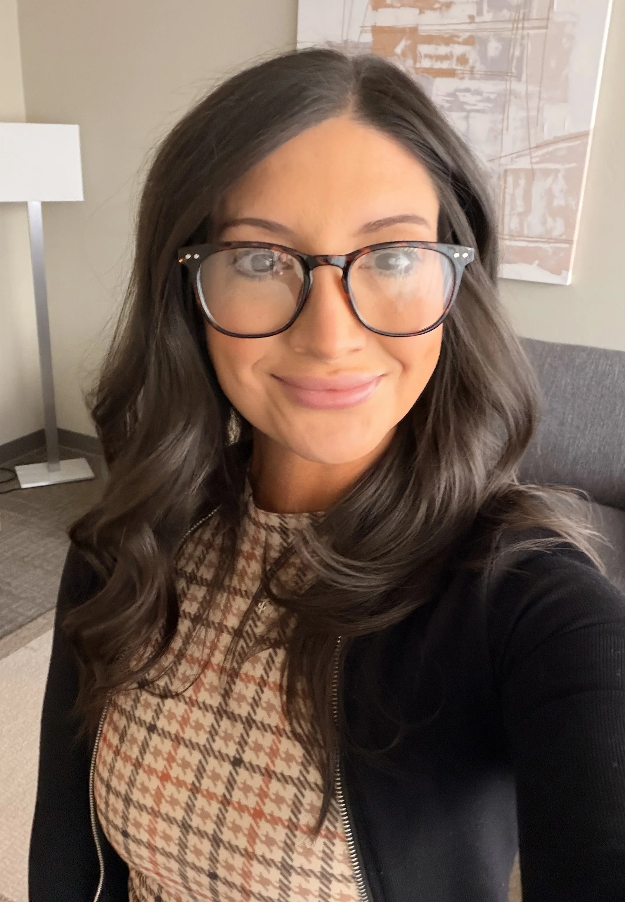 A woman with long, wavy brown hair and glasses, smiling, taking a selfie indoors. She is wearing a patterned top and a black jacket. There is a lamp and a wall art in the background.