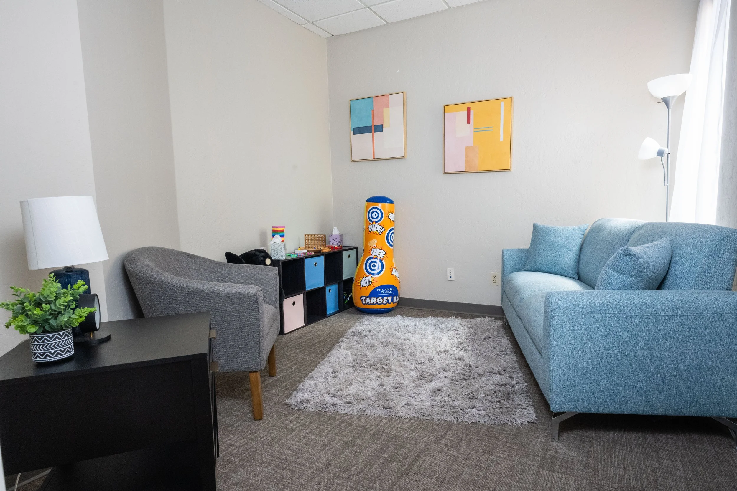 A cozy room with blue and gray furniture, colorful modern paintings on the wall, a small black storage unit, an inflatable punching bag, a shaggy rug, a black side table with green plant, and a standing lamps near a window with white curtains.