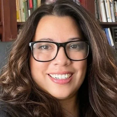 A woman with long brown hair, wearing glasses, smiling in front of a bookshelf.