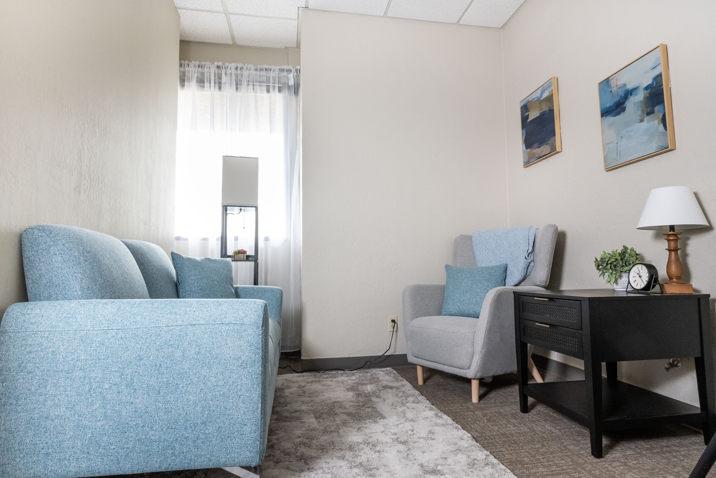 Therapy room with light blue sofa, gray armchair with a blue pillow, black side table with a white clock, a wooden lamp, plants, and framed artwork on the wall. A window with sheer curtains lets in natural light.