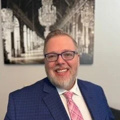A man with glasses, a beard, and light brown hair smiling, wearing a blue suit and pink tie, in front of a black and white cityscape photograph with chandeliers