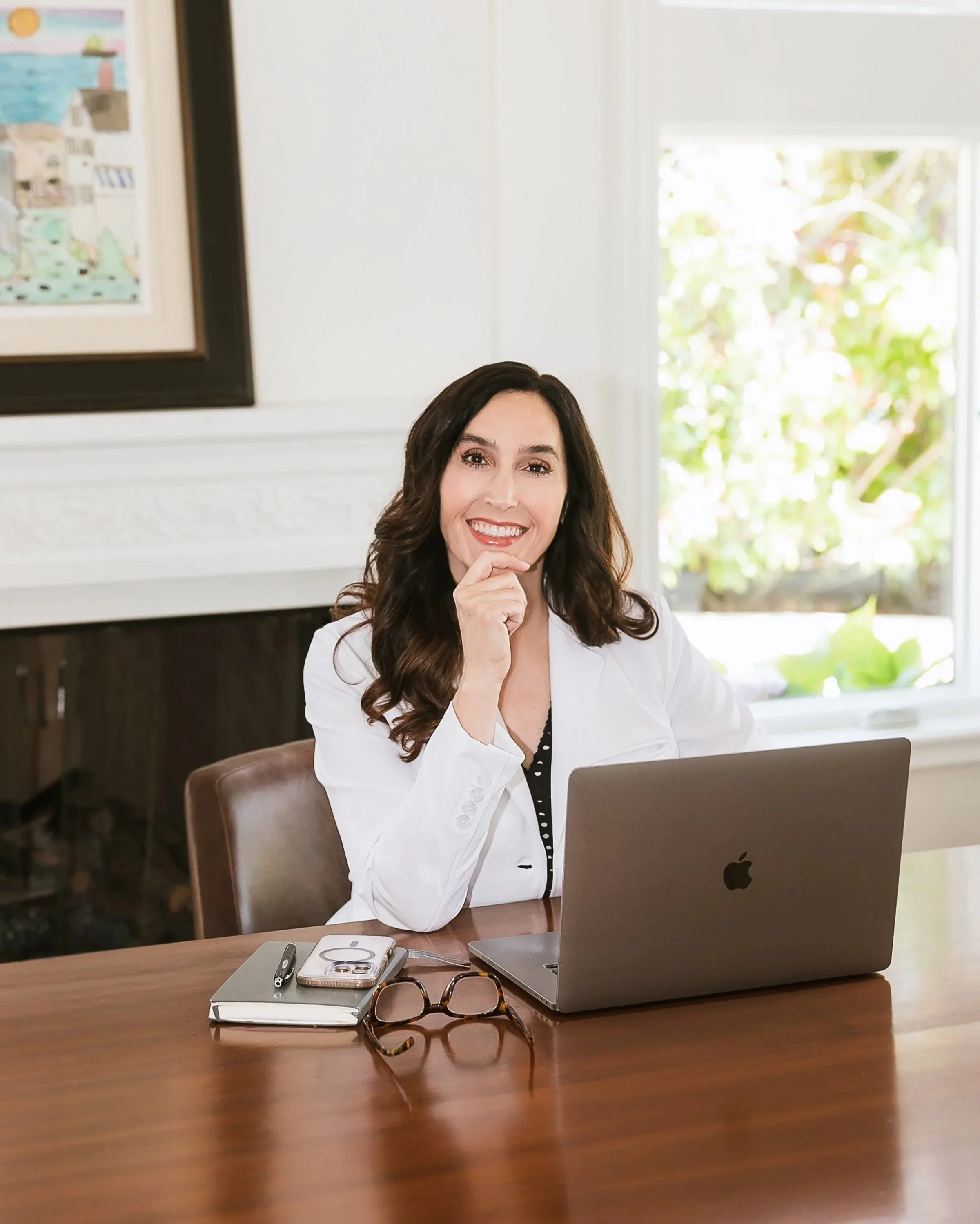 Dr. Vinther smiling and sitting at a desk with a laptop, smartphone, notebook, pair of glasses, and a pen. She has dark, wavy hair and is wearing a white blazer.