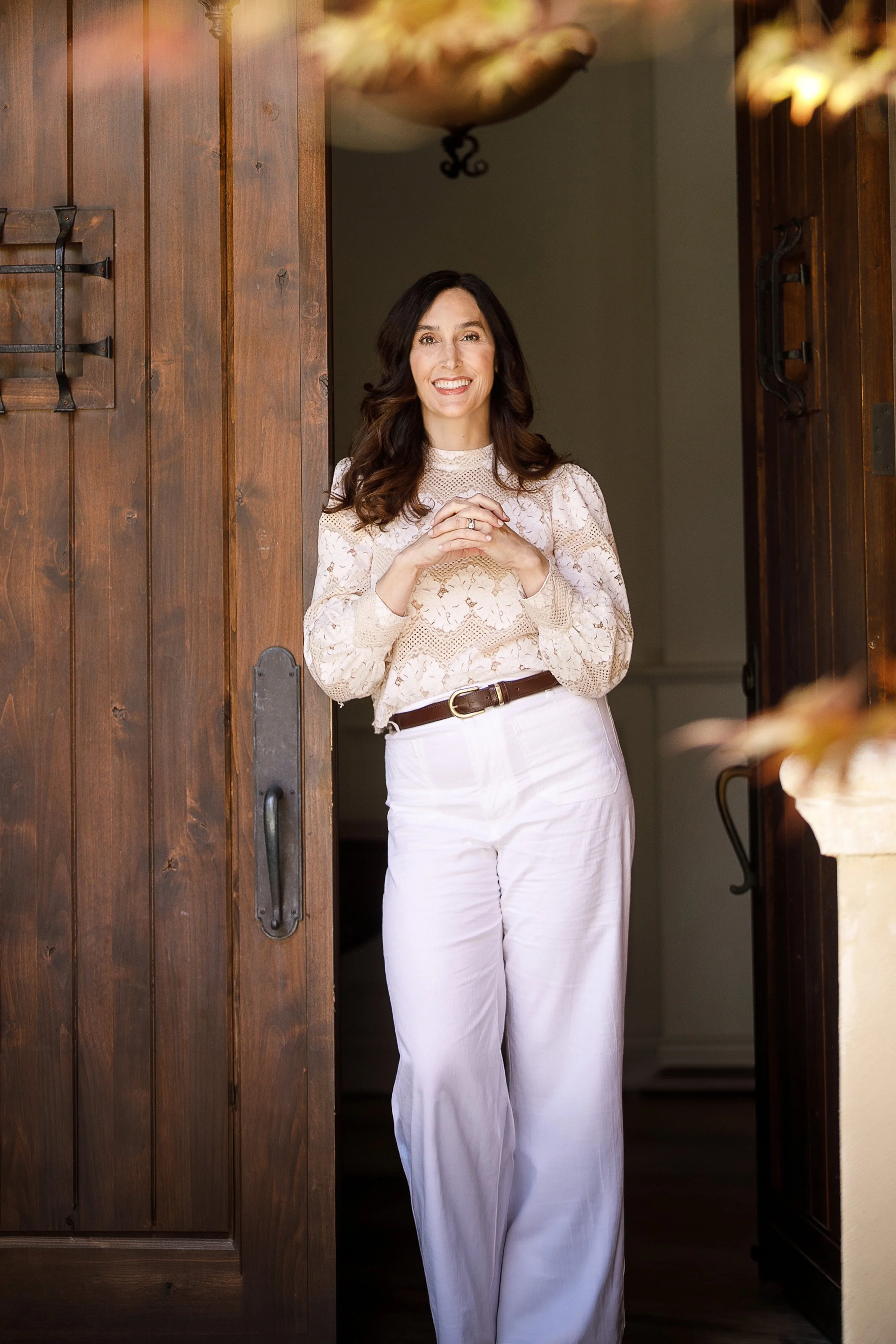 Dr. Vinther, wearing a cream lace blouse and white pants, standing at a wooden doorway, smiling.