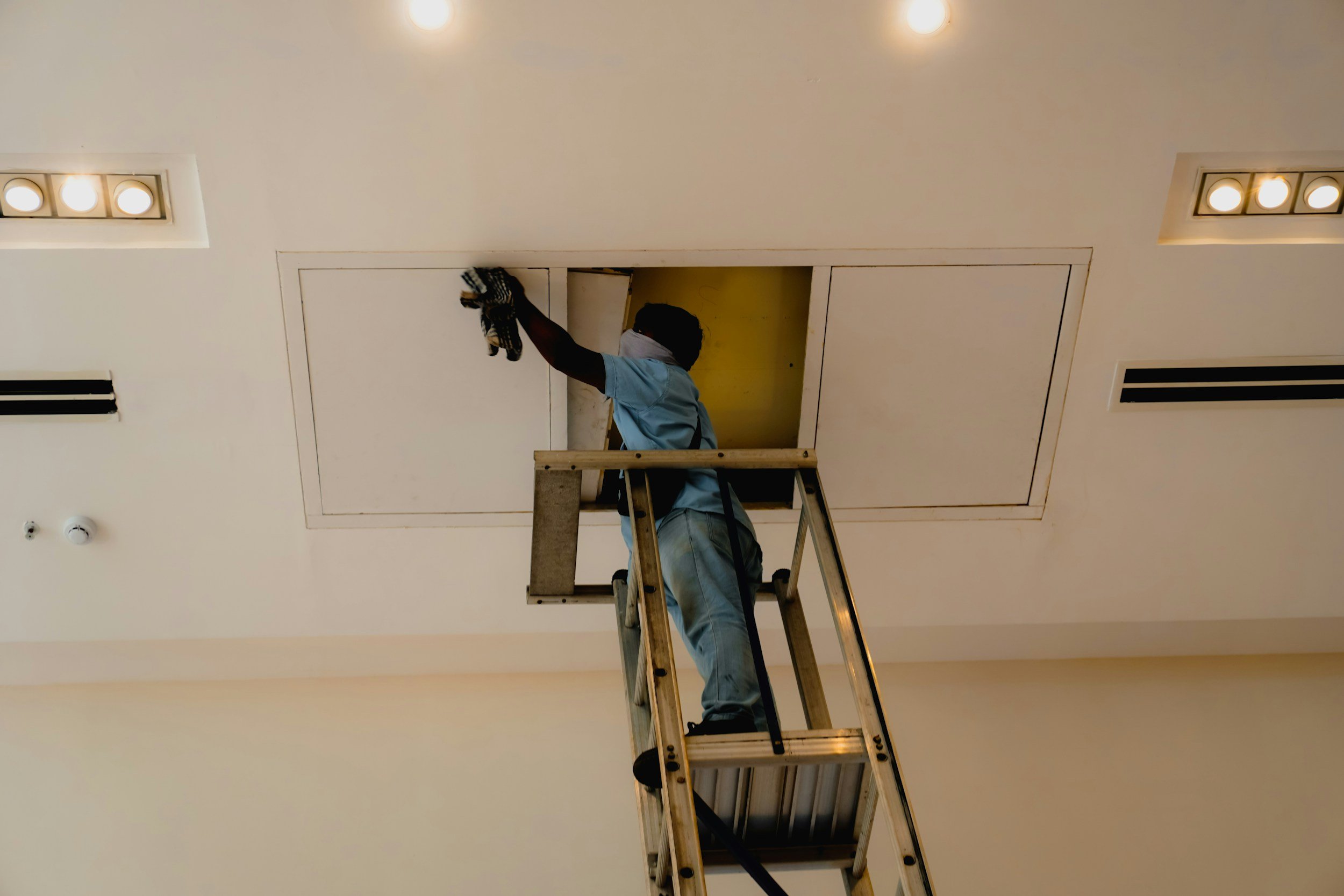 A worker is on a ladder installing or repairing a ceiling panel in a white ceiling room. The worker is wearing a light blue shirt and gloves, and has a towel around their neck.