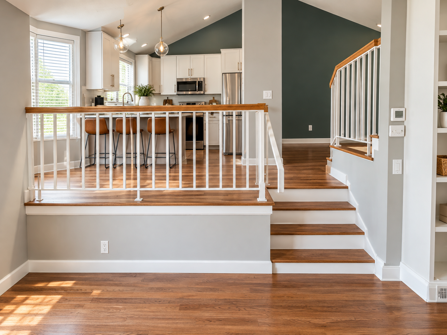 A modern kitchen and living space with hardwood floors, white cabinets, stainless steel appliances, bar stools at a kitchen island, and a staircase with white railing leading to a higher level.