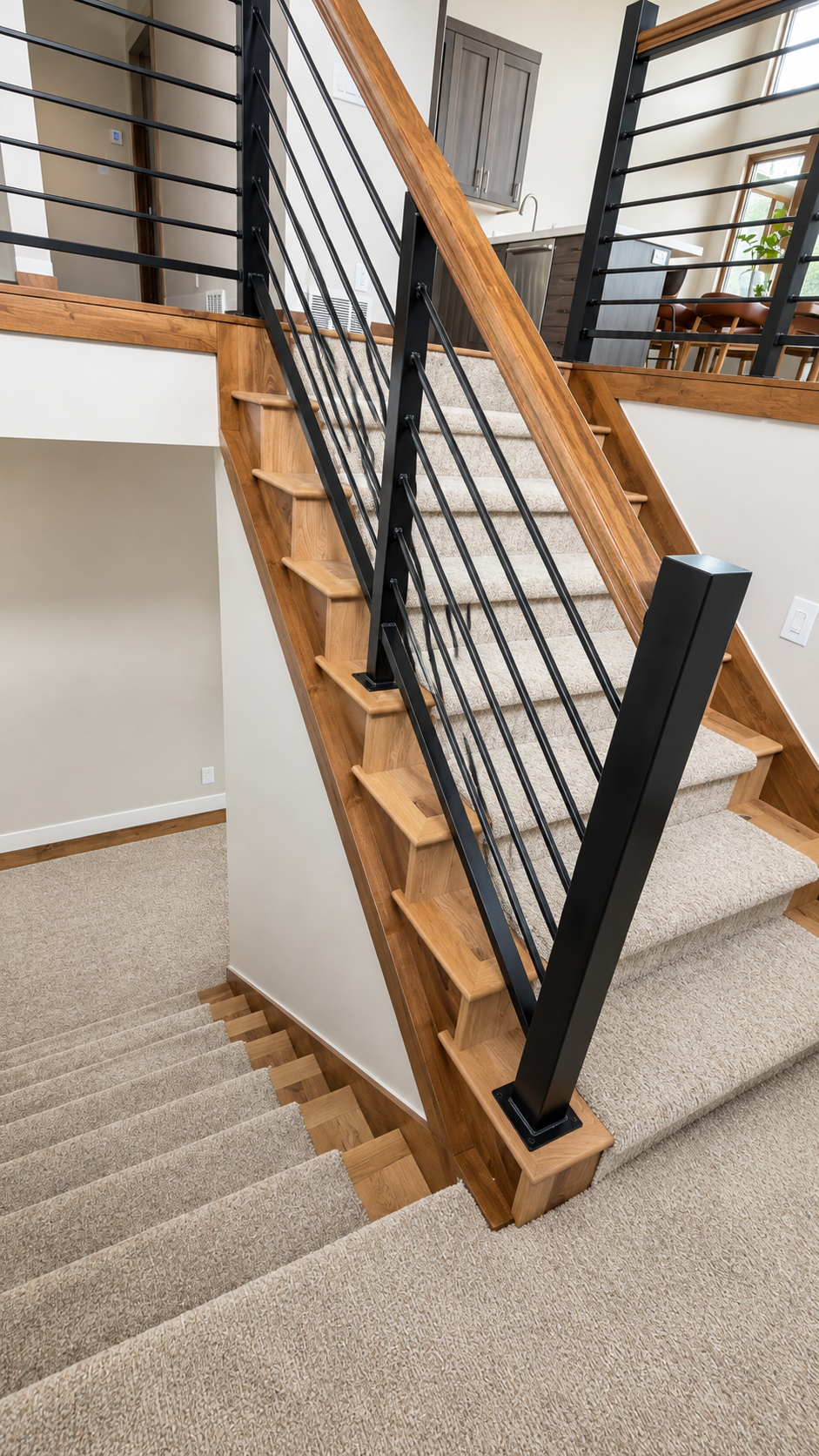 Interior of a modern staircase with wooden steps, a black metal railing, and beige carpeted stairs leading to upstairs, with part of a kitchen and dining area visible in the background.