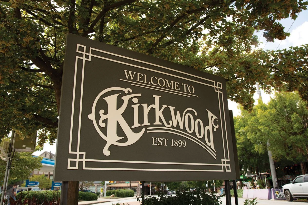 Welcome to Kirkwood sign in a park-like setting, with trees and buildings in the background.