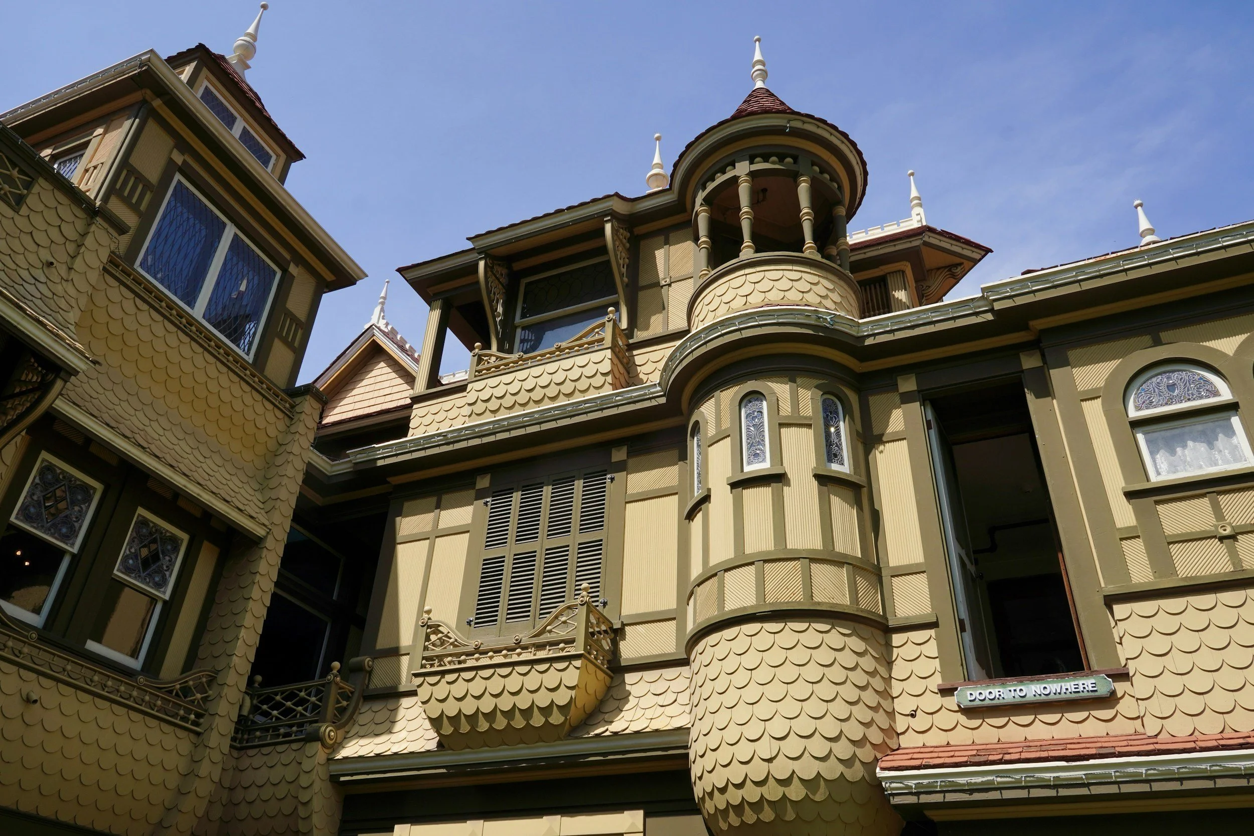 A large, ornate Victorian-style house with beige siding, decorative shingles, multiple turrets topped with spires, and stained glass windows. A sign near the bottom reads 'Door to Nowhere'.