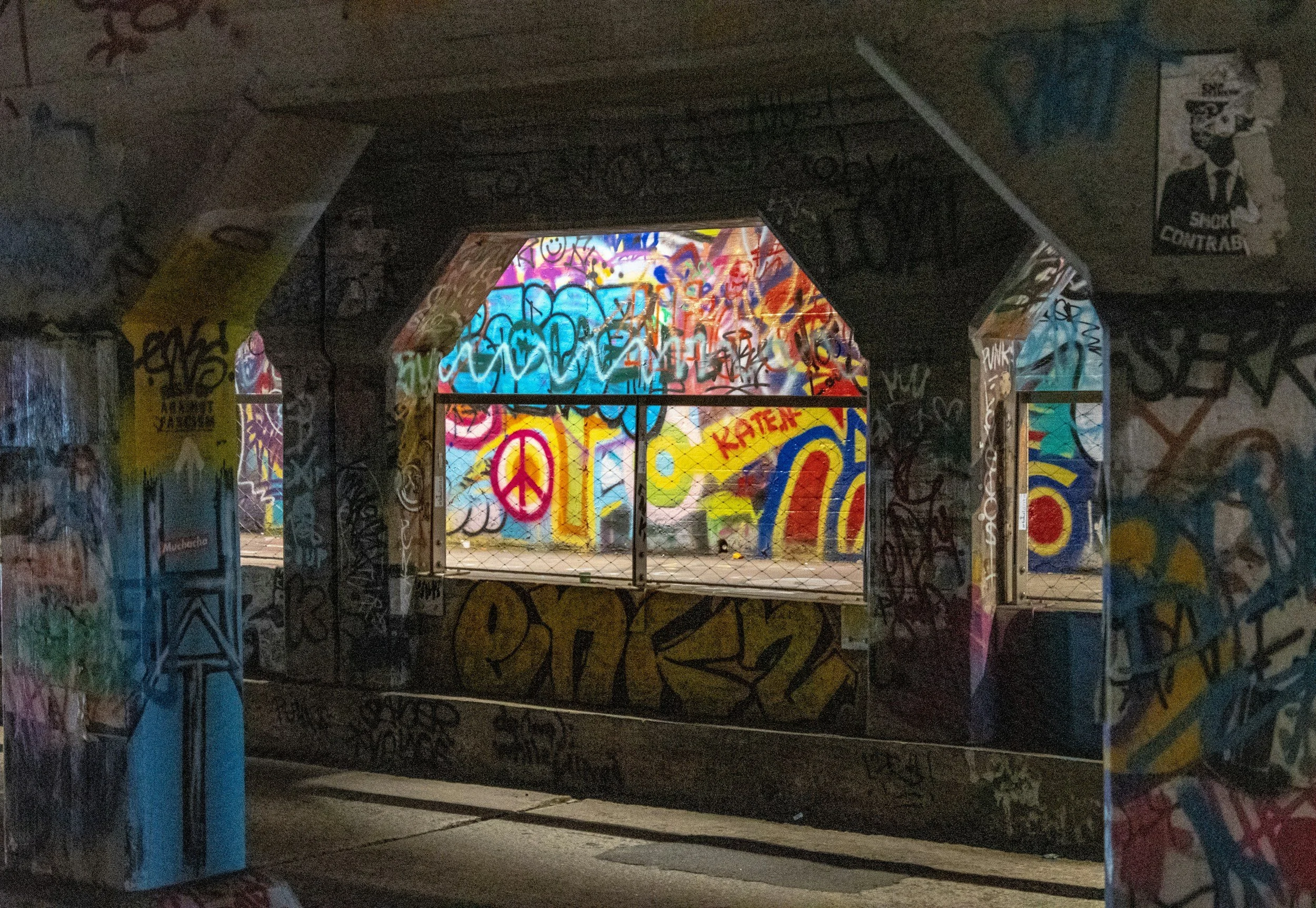 Graffiti-covered underpass with bright, colorful street art and a chain-link fence, illuminated by ambient light at night.