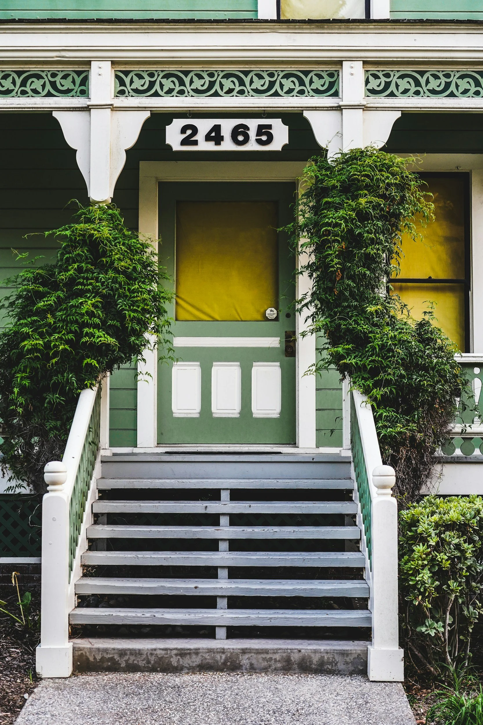 Front porch of a house with a set of stairs, green door with yellow window covering, white railing, and climbing plants on both sides, house number 2465 displayed above the door.