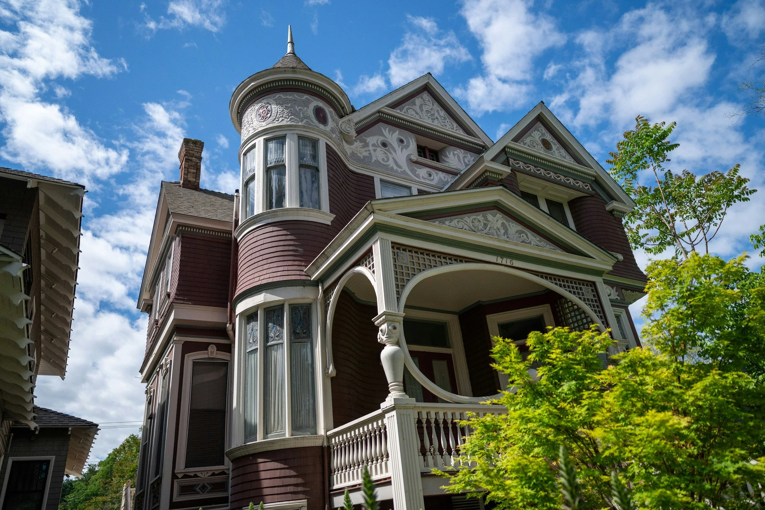 A Victorian-style house with a turret and wraparound porch, featuring ornate woodwork and decorative trim, against a blue sky with clouds and some green trees.