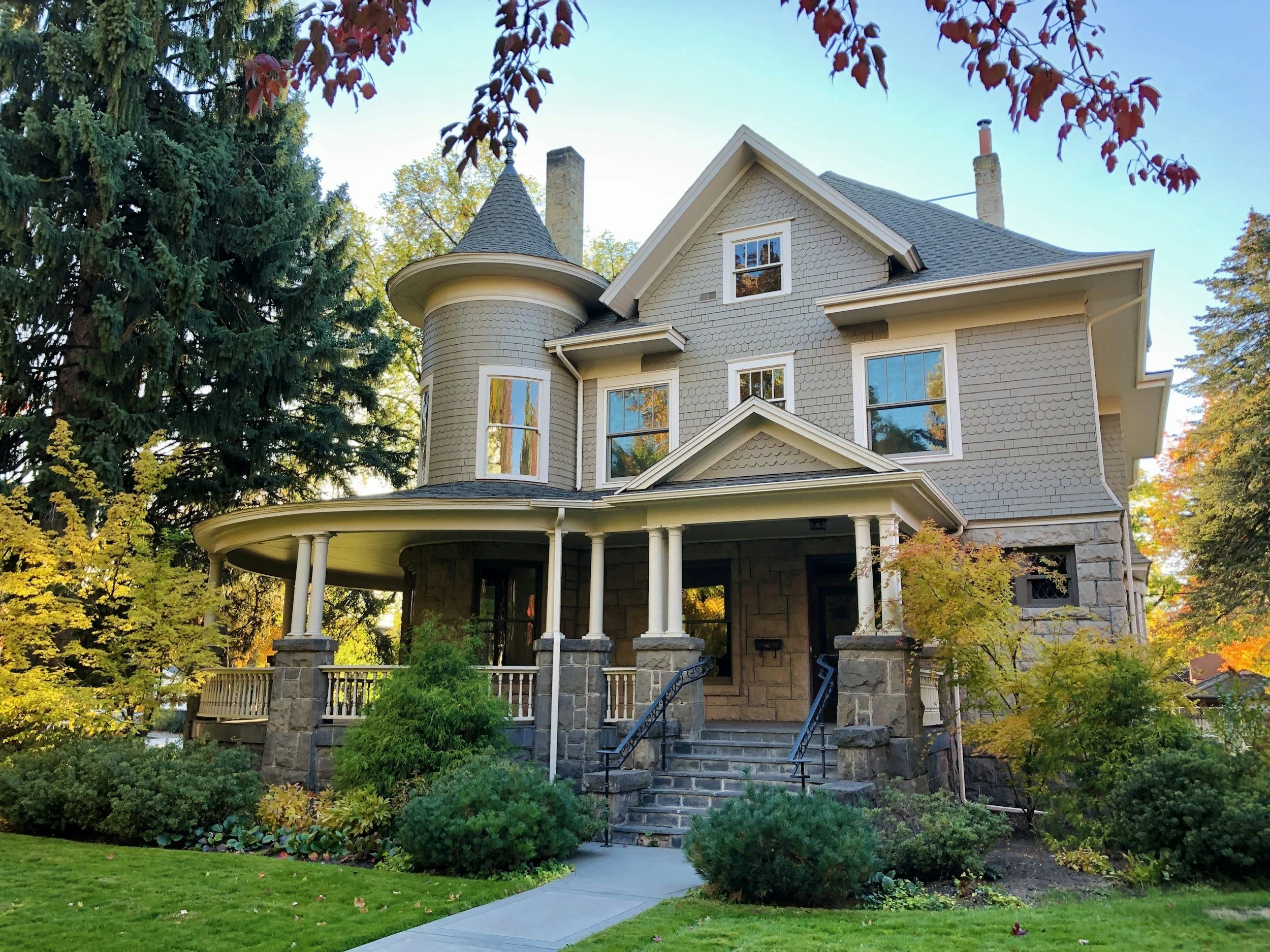 A light gray Victorian-style house with a turret, bay windows, and a porch surrounded by trees with fall foliage, with a well-maintained lawn and stone steps leading to the front door.