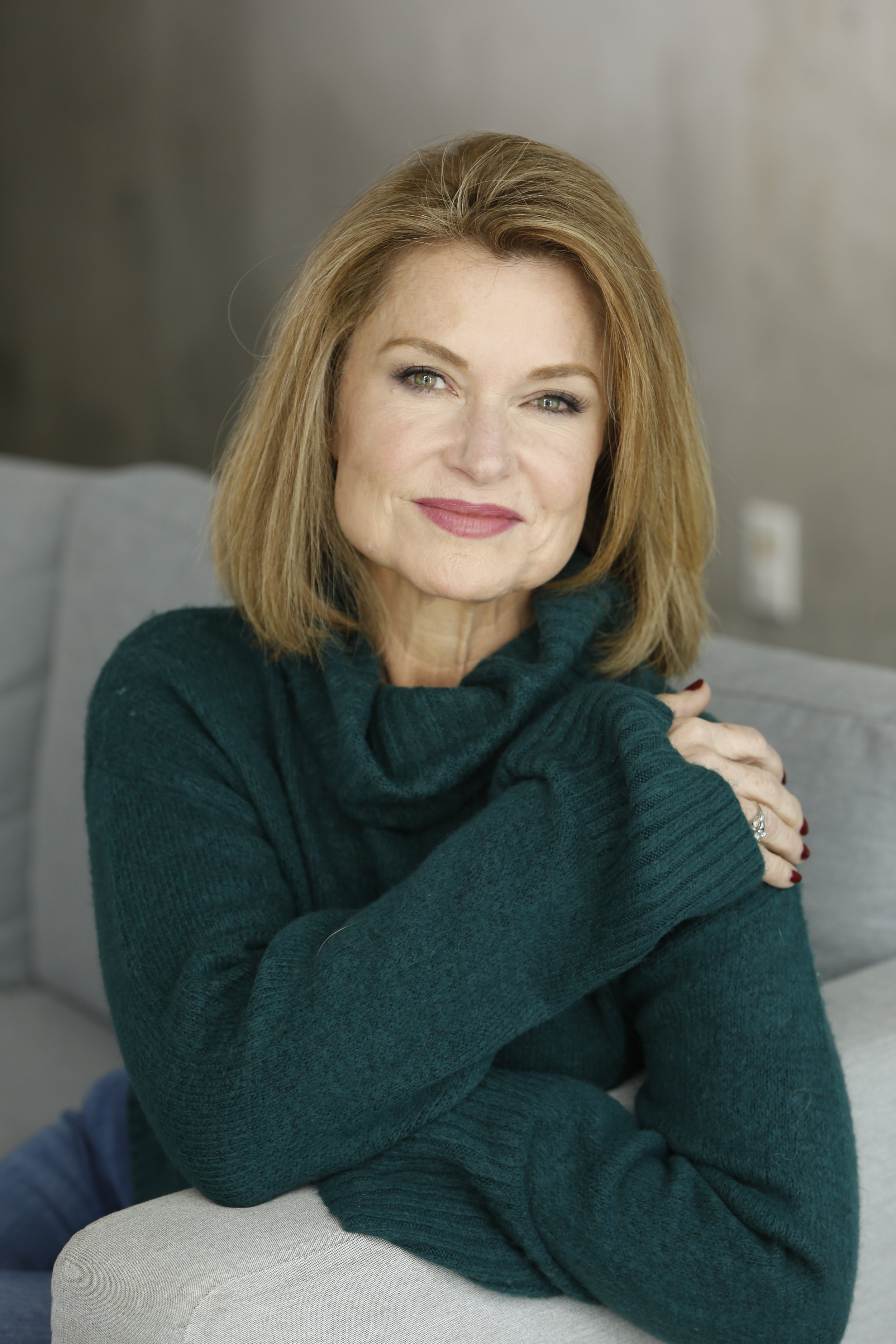 A woman with shoulder-length red hair, green eyes, and fair skin, wearing a dark green turtleneck sweater, smiling and sitting on a light gray chair, with a blurred background indoors.