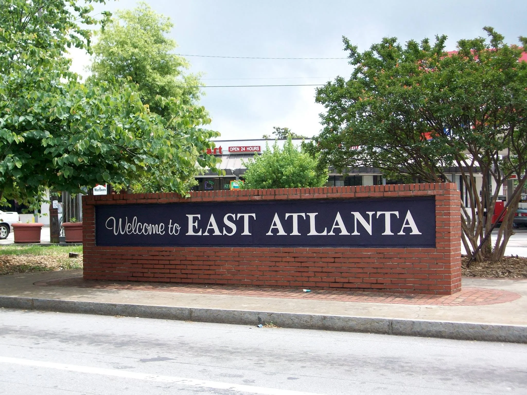 Welcome to East Atlanta sign on a brick wall, with lush green trees behind it and a clear sky overhead.