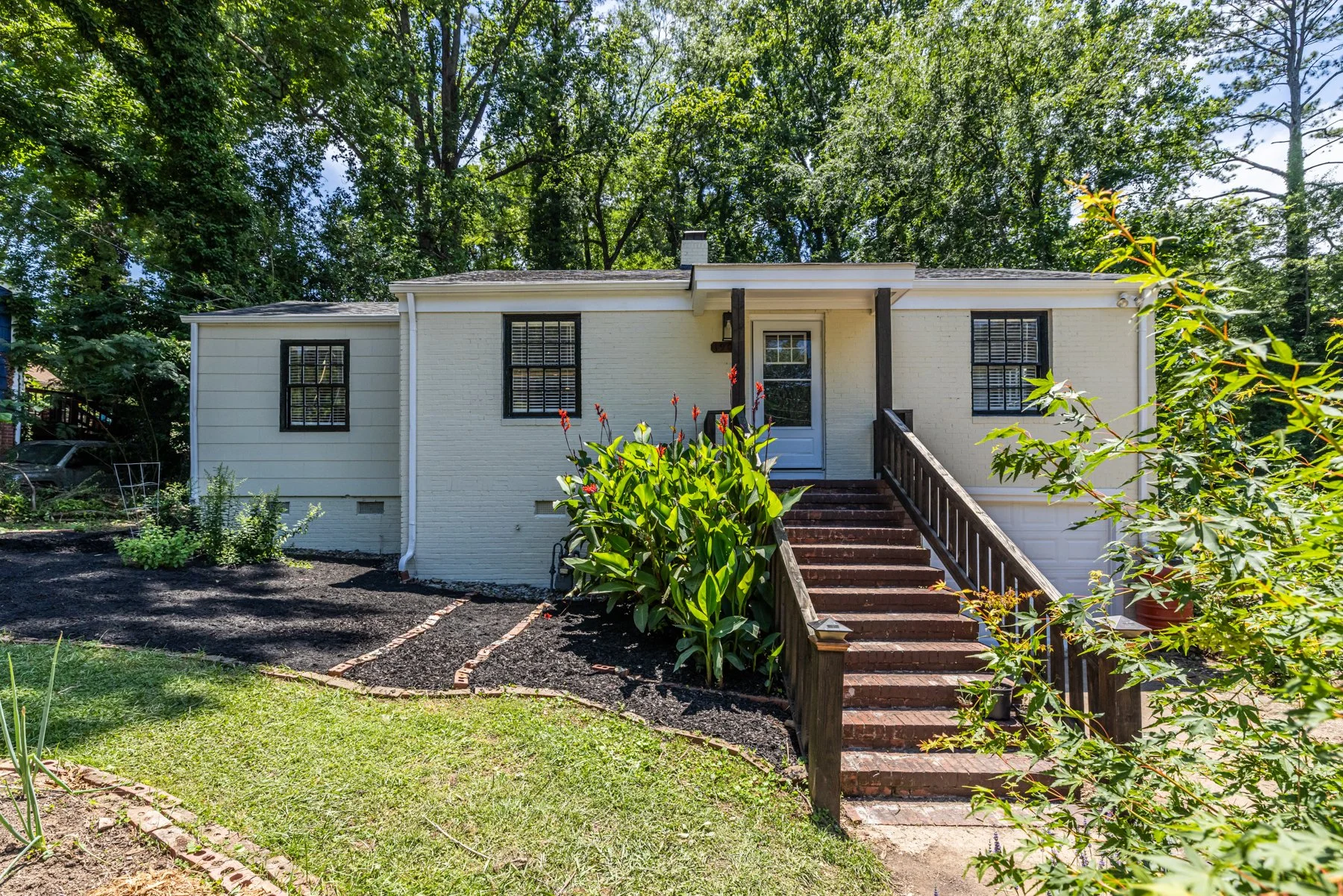 White brick house with black window shutters and stairs leading to front door, surrounded by green trees and garden plants.