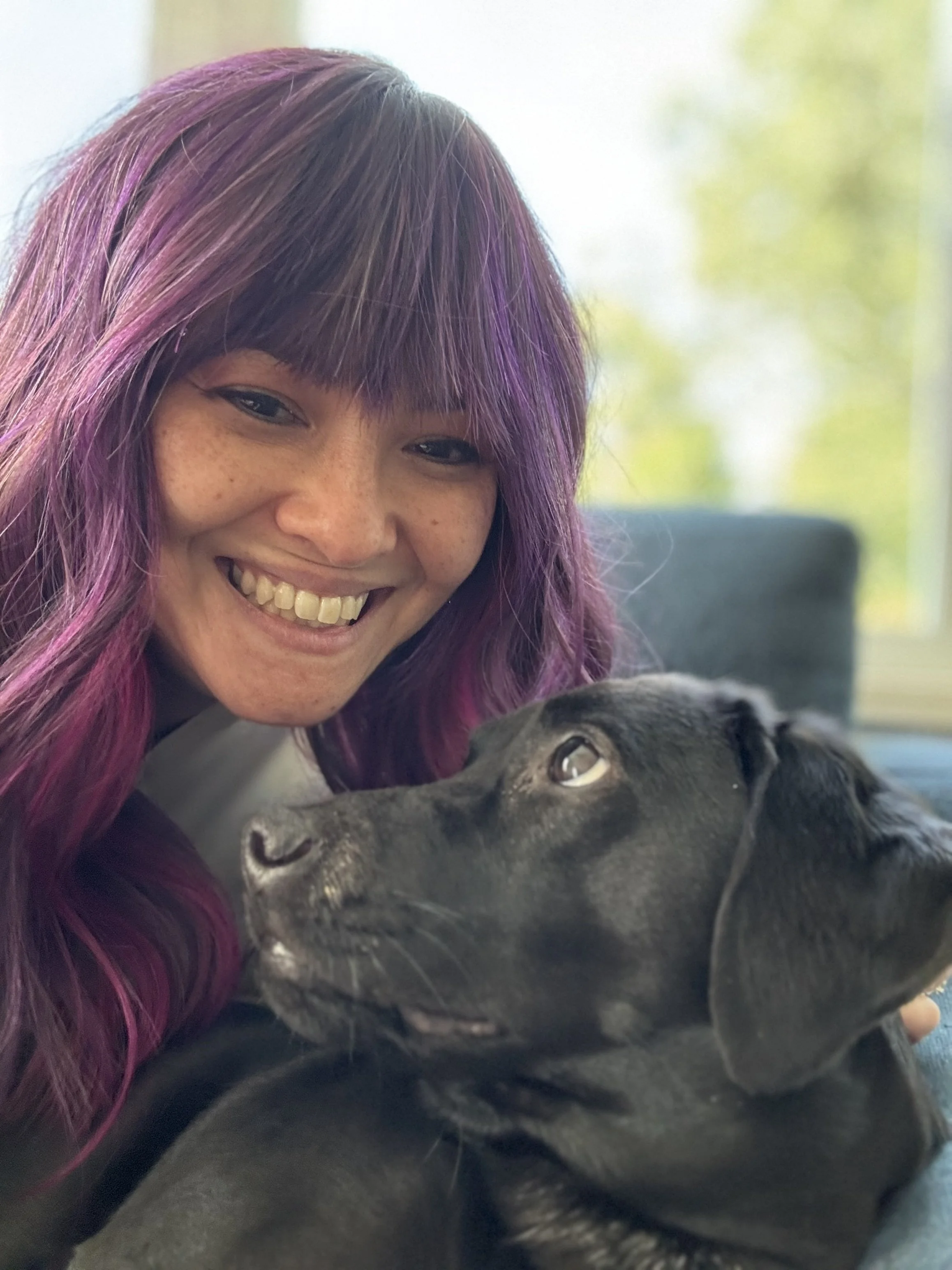 A woman with purple hair smiling and holding a black dog, indoors with a blurred window and natural light in the background.