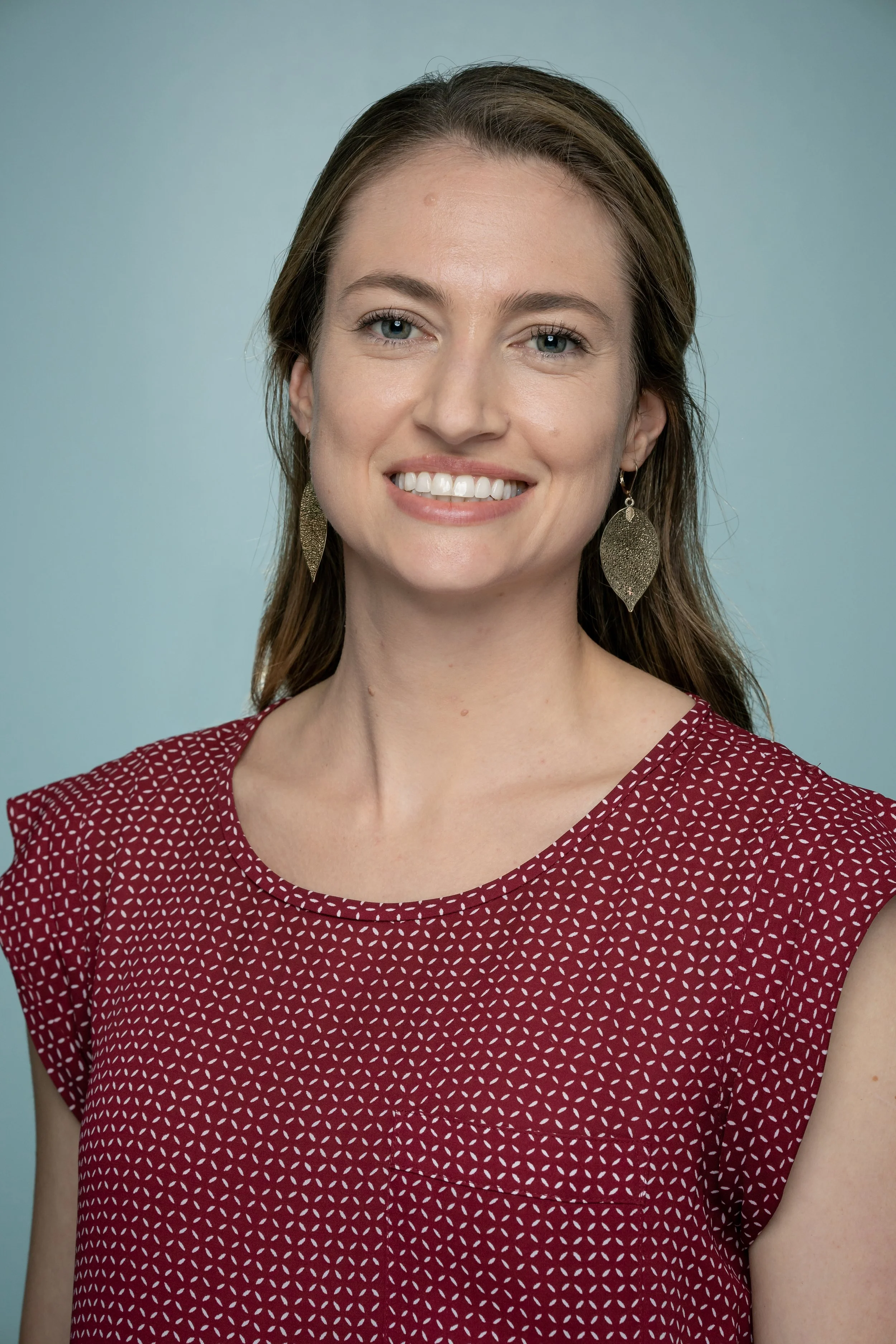 Head-and-shoulders portrait of a young woman with long brown hair, blue eyes, and earrings, smiling in a maroon polka dot top against a light blue background.