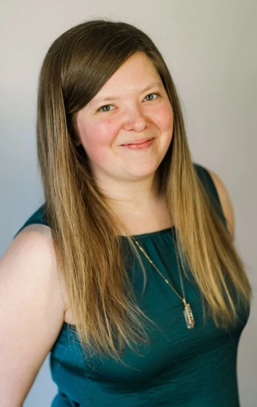 A young woman with long, light brown hair and blue eyes, smiling at the camera, wearing a dark teal sleeveless dress and a gold necklace with a rectangular pendant, standing against a plain, light-colored background.