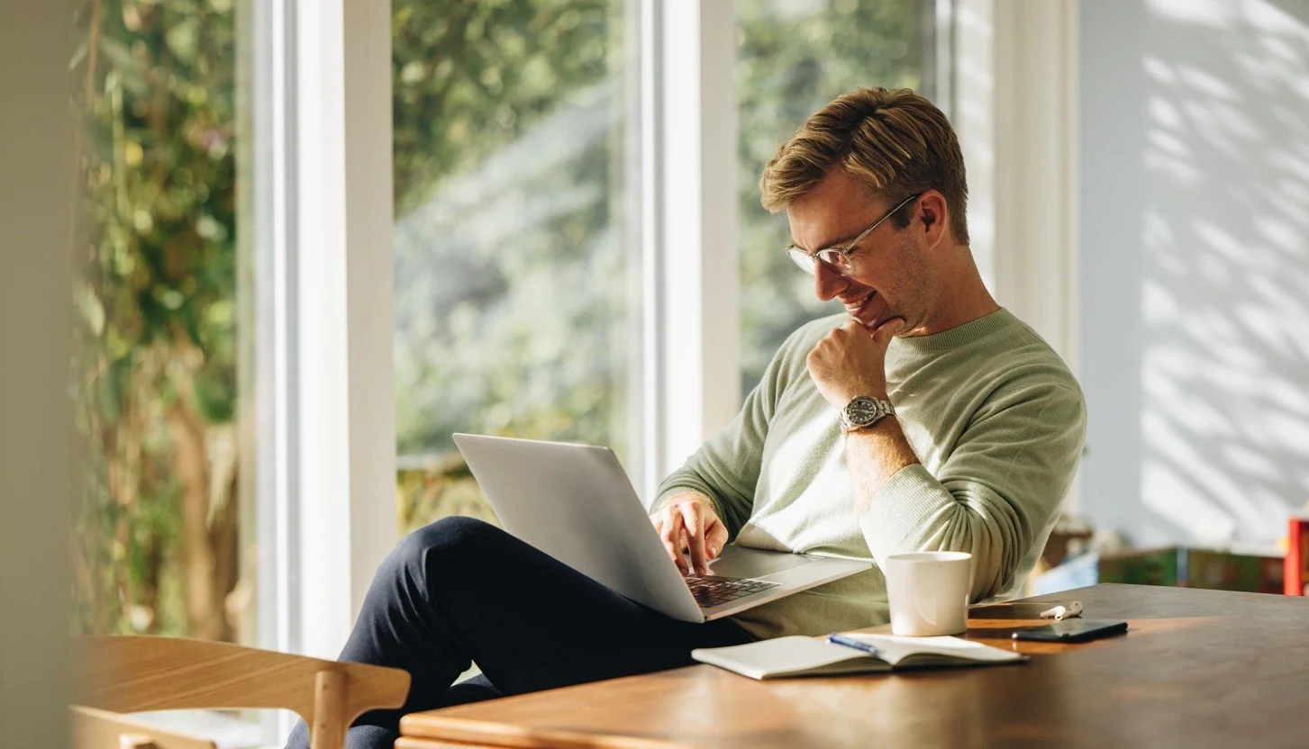 A young man with glasses in a light-colored sweater, sitting at a wooden table near large windows, smiling while looking at his laptop, with a coffee mug, notebook, and pen on the table.