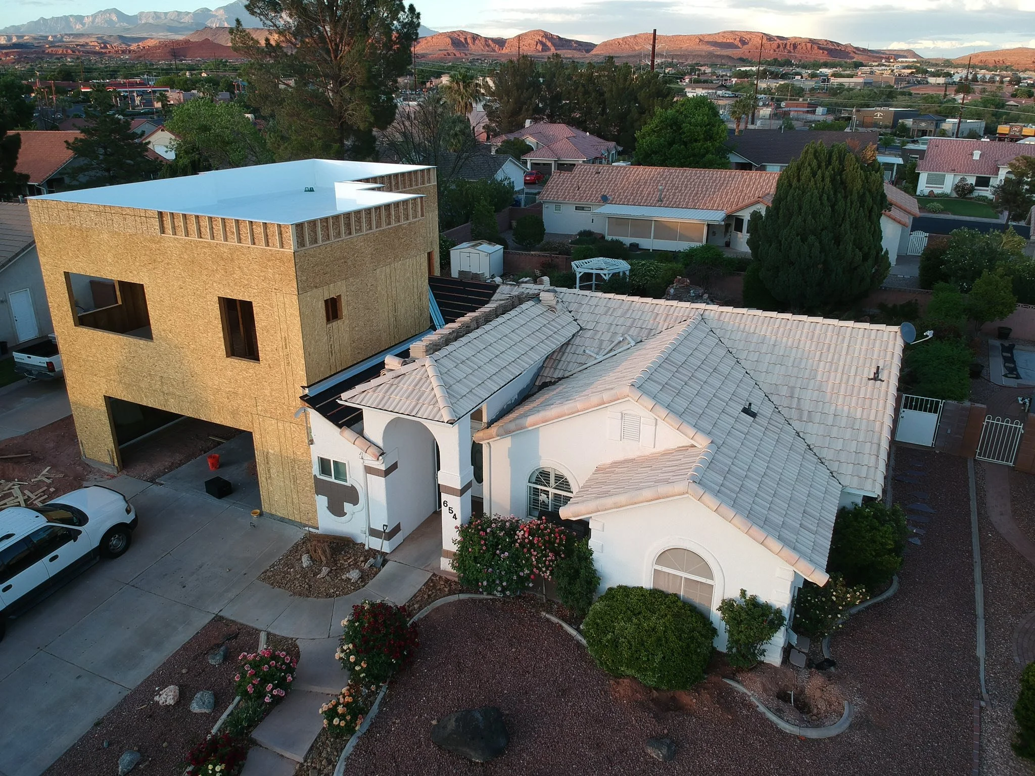 Barlovento en un vecindario con casas de techos de teja y un edificio en construcción en primer plano, rodeado de árboles flores y aceras, con montañas en el fondo y una tarde soleada.