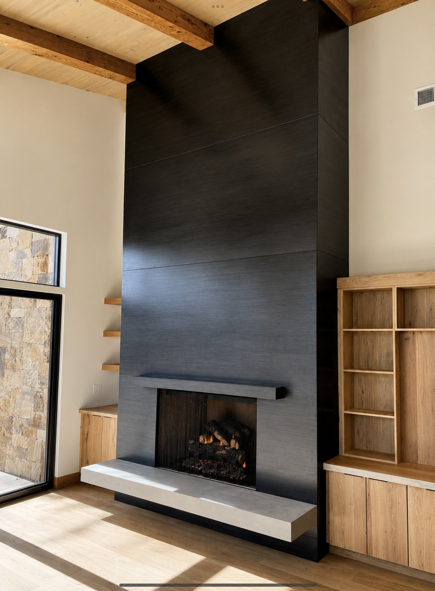 Modern living room with a black and gray fireplace, wooden ceiling beams, a brick wall, and light wood flooring.
