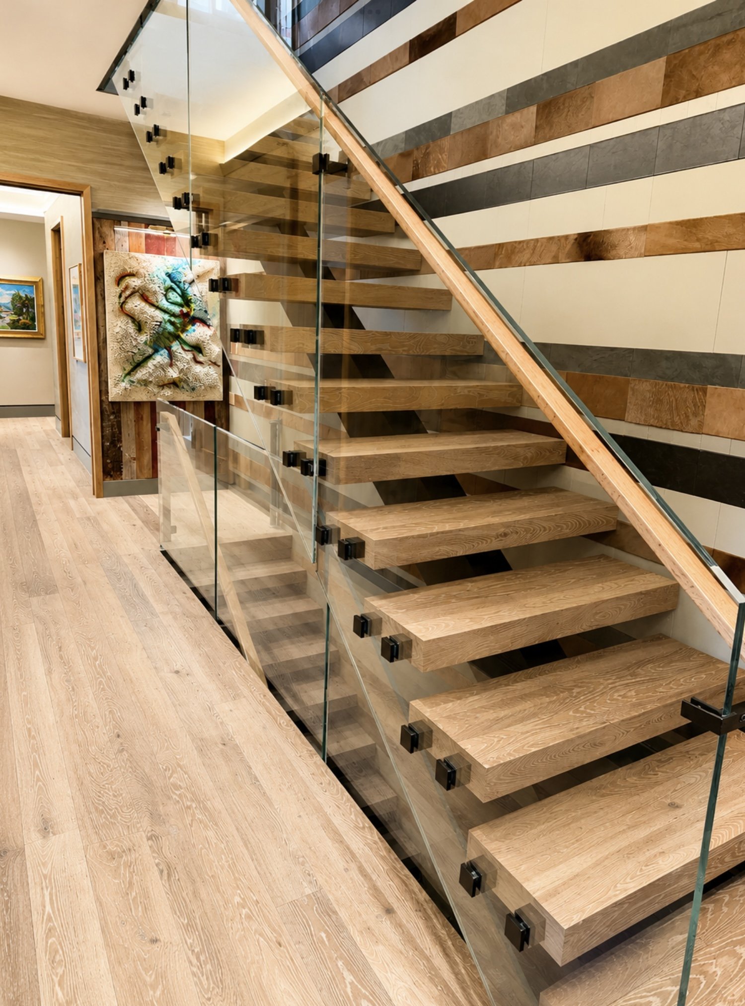 Interior view of a modern staircase with wooden steps, glass railing, and wall decor, in a well-lit building with wooden flooring and artwork on the walls.