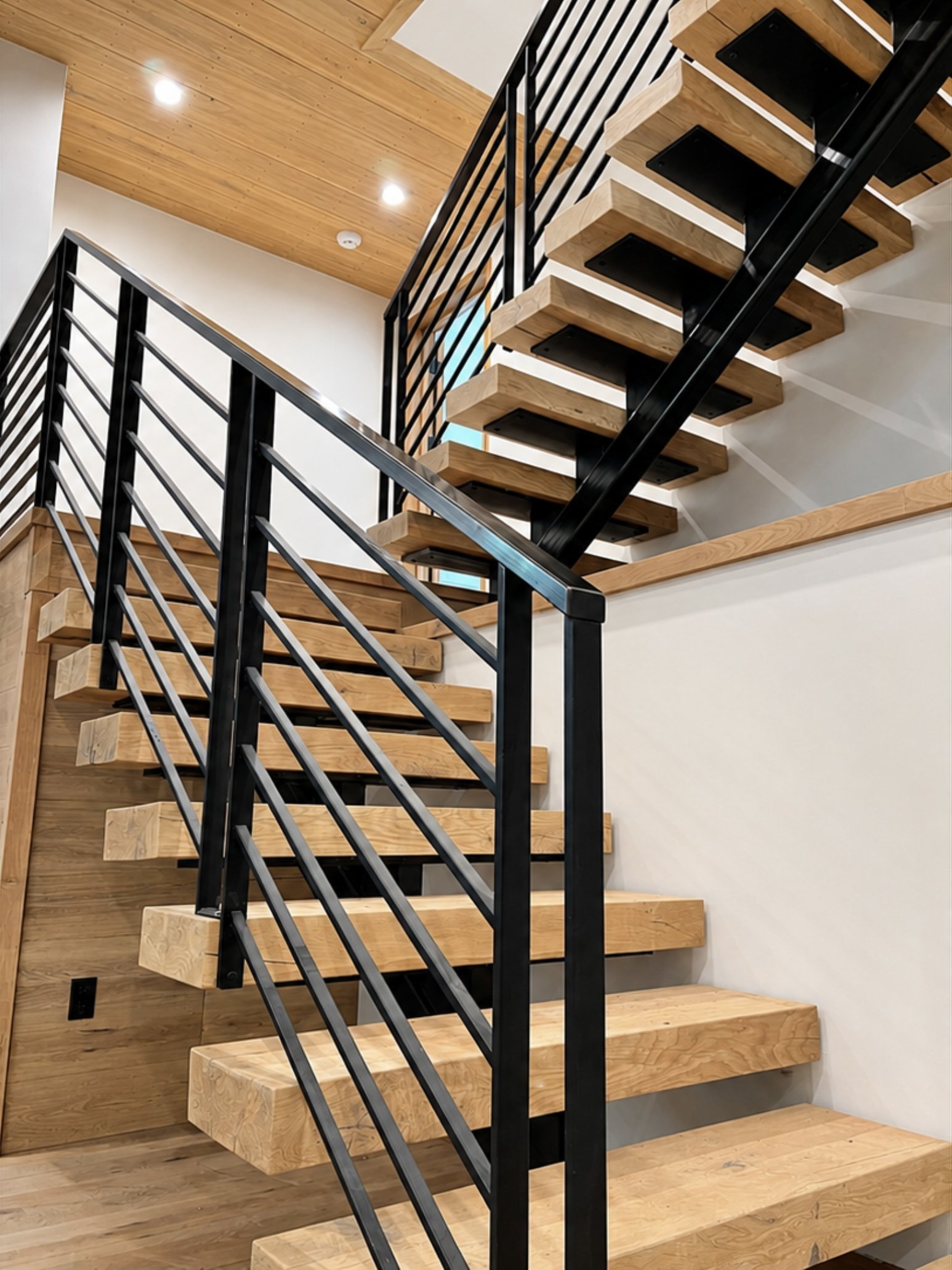 Interior view of a modern staircase with wooden steps and black metal railings, leading to the upper floor. The ceiling above the staircase is wooden with recessed lighting, and the walls are painted white.