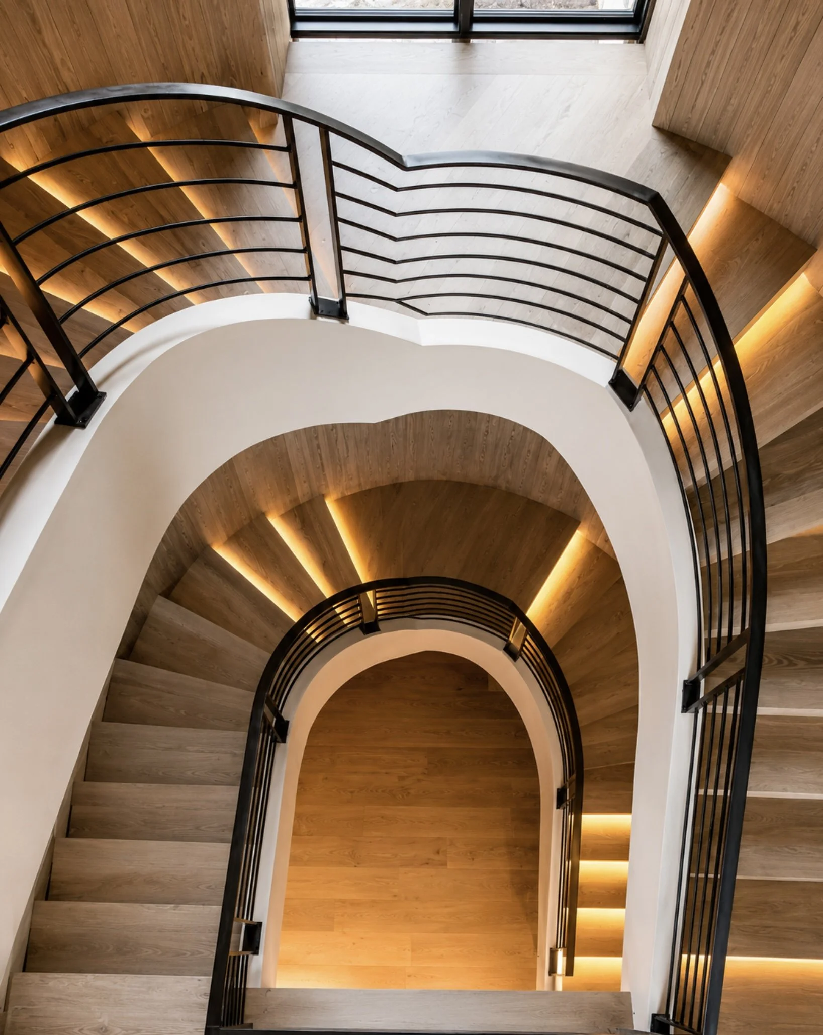 A view from the top of a spiral staircase with wooden steps, black metal handrails, and built-in lighting along the walls, looking down.