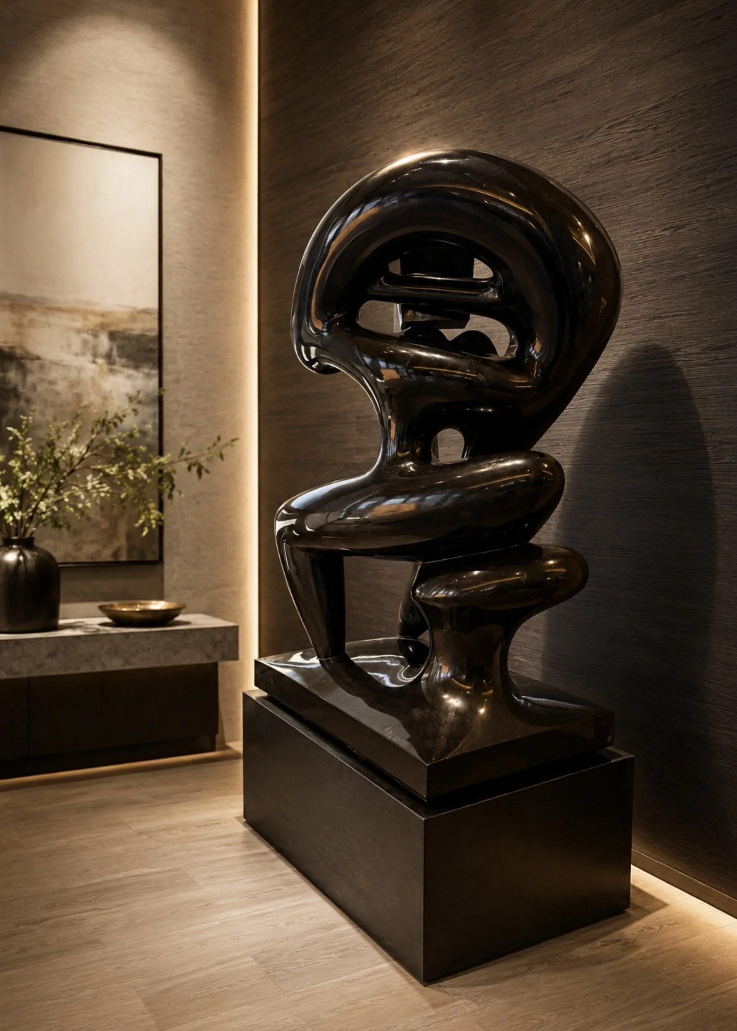 A shiny black, abstract sculpture on a black pedestal in a modern interior space with wood flooring, a dark wooden wall, and a small table with a vase of flowers and a decorative dish.