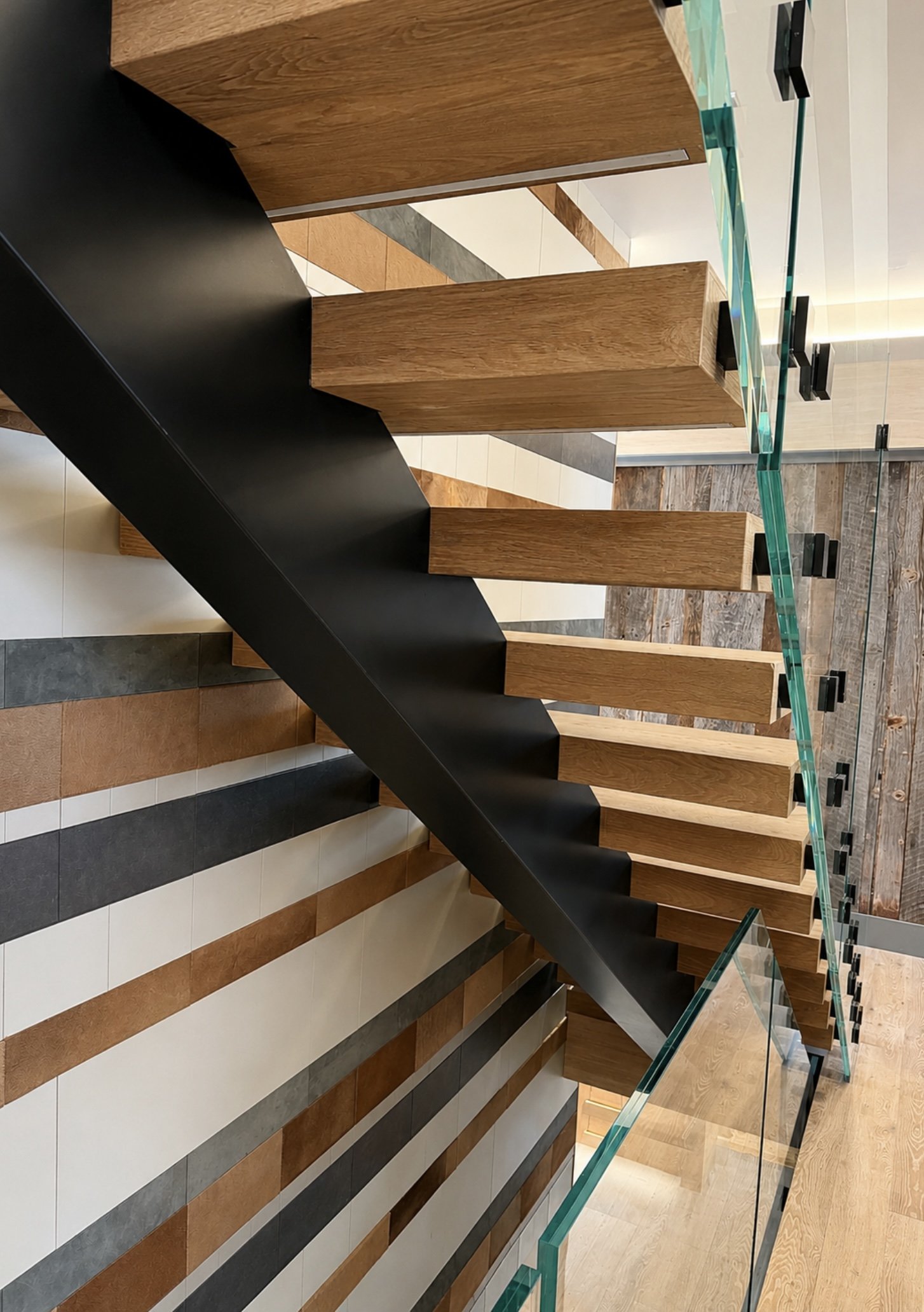 Interior view of a modern staircase with wooden steps, a black side panel, and a glass railing, in a building with striped tile walls and wooden flooring.