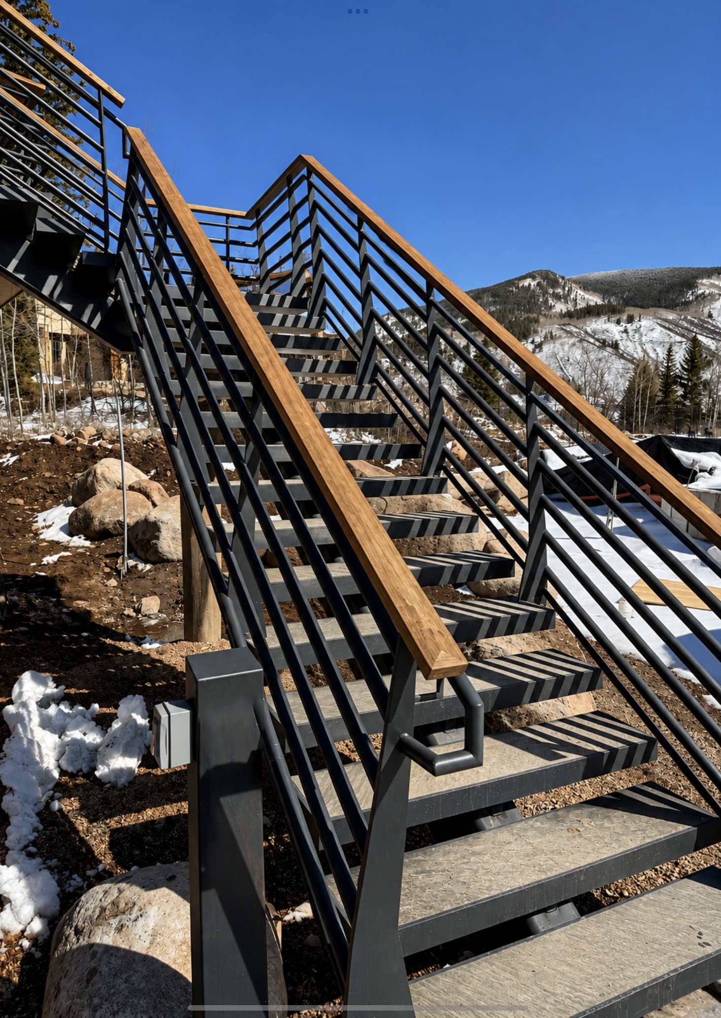 Metal stairs with wooden handrails installed outdoors on a construction site with mountains and snow in the background.