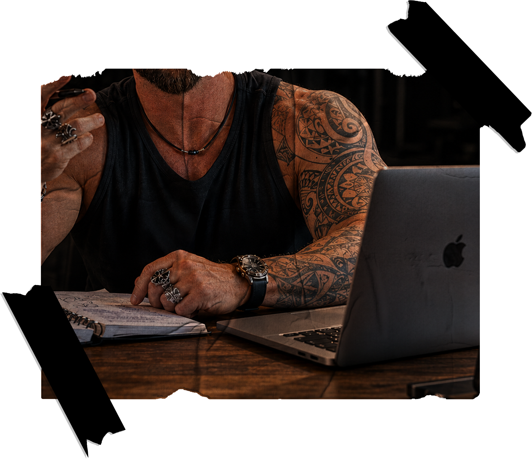 A man with tattoos on his arms, wearing rings, a watch, and necklaces, sitting at a wooden table with a laptop and a notebook, holding a smartphone.