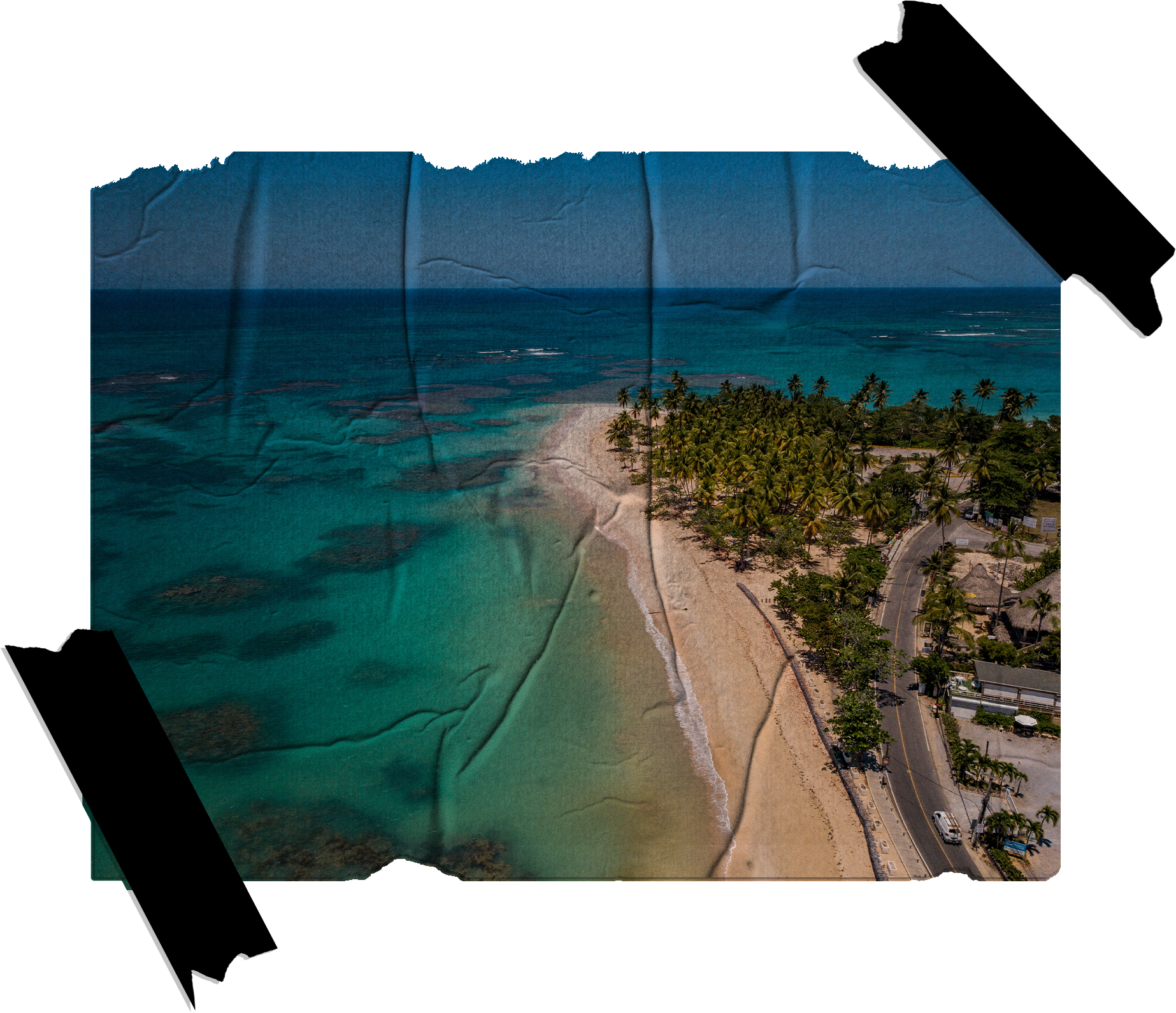 Aerial view of a tropical beach with turquoise water, white sand, and palm trees, adjacent to a road with parked cars and buildings.