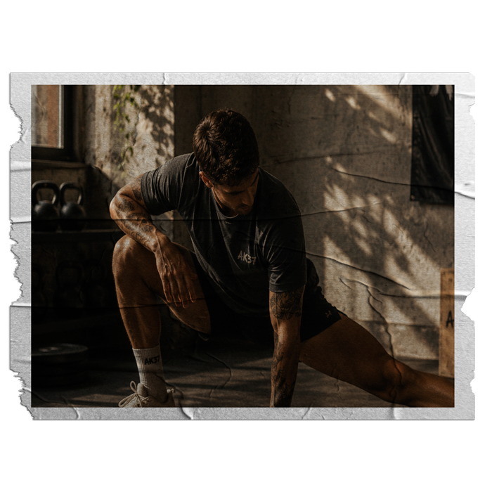 A man in a black t-shirt and shorts stretching on the floor of a gym with sunlight streaming through the window.