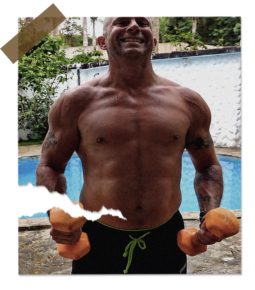 A shirtless man standing outdoors by a swimming pool, holding two orange dumbbells, smiling, with tattoos on his arms.
