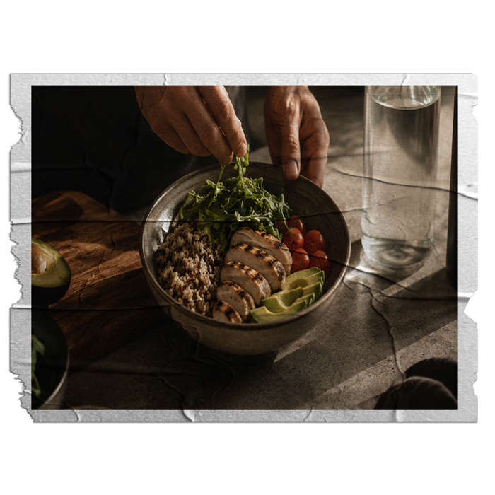 A person preparing a bowl of salad with grilled chicken, grape tomatoes, lime wedges, quinoa, and leafy greens on a table next to a glass of water.